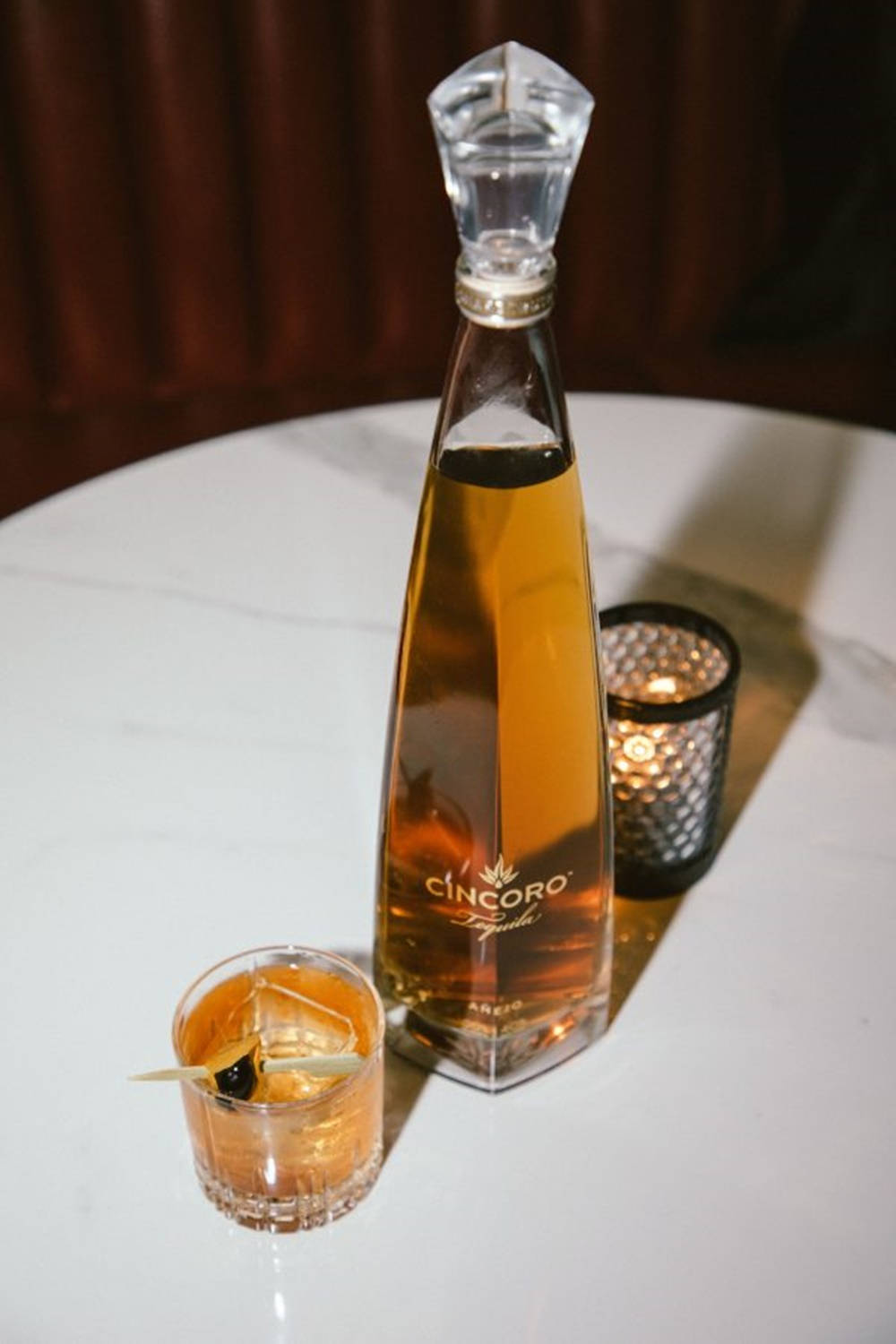 bottle 桌布 壁紙 — 一個時尚的Cincoro龍舌蘭酒瓶和雞尾酒杯放在白色桌子上，旁邊有一根在溫暖環境燈光下閃爍的蠟燭。