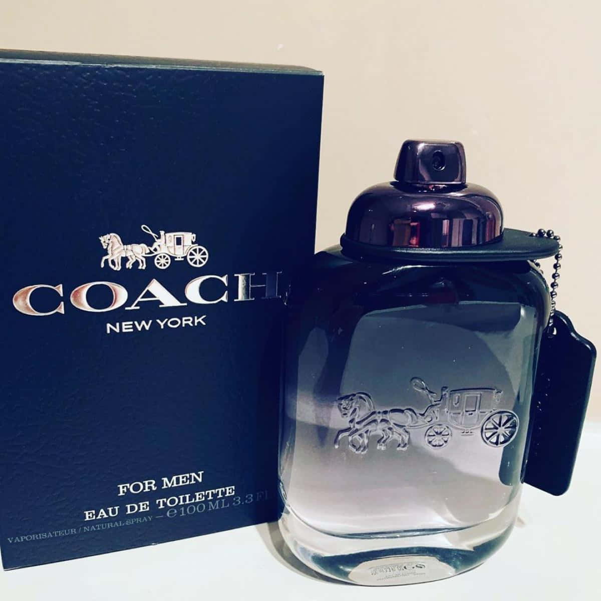 Coach 桌布 壁紙 — 一個時尚的Coach for men男士香水瓶，帶有浮雕馬車圖案，旁邊是其深藍色品牌盒子，放在白色表面上。