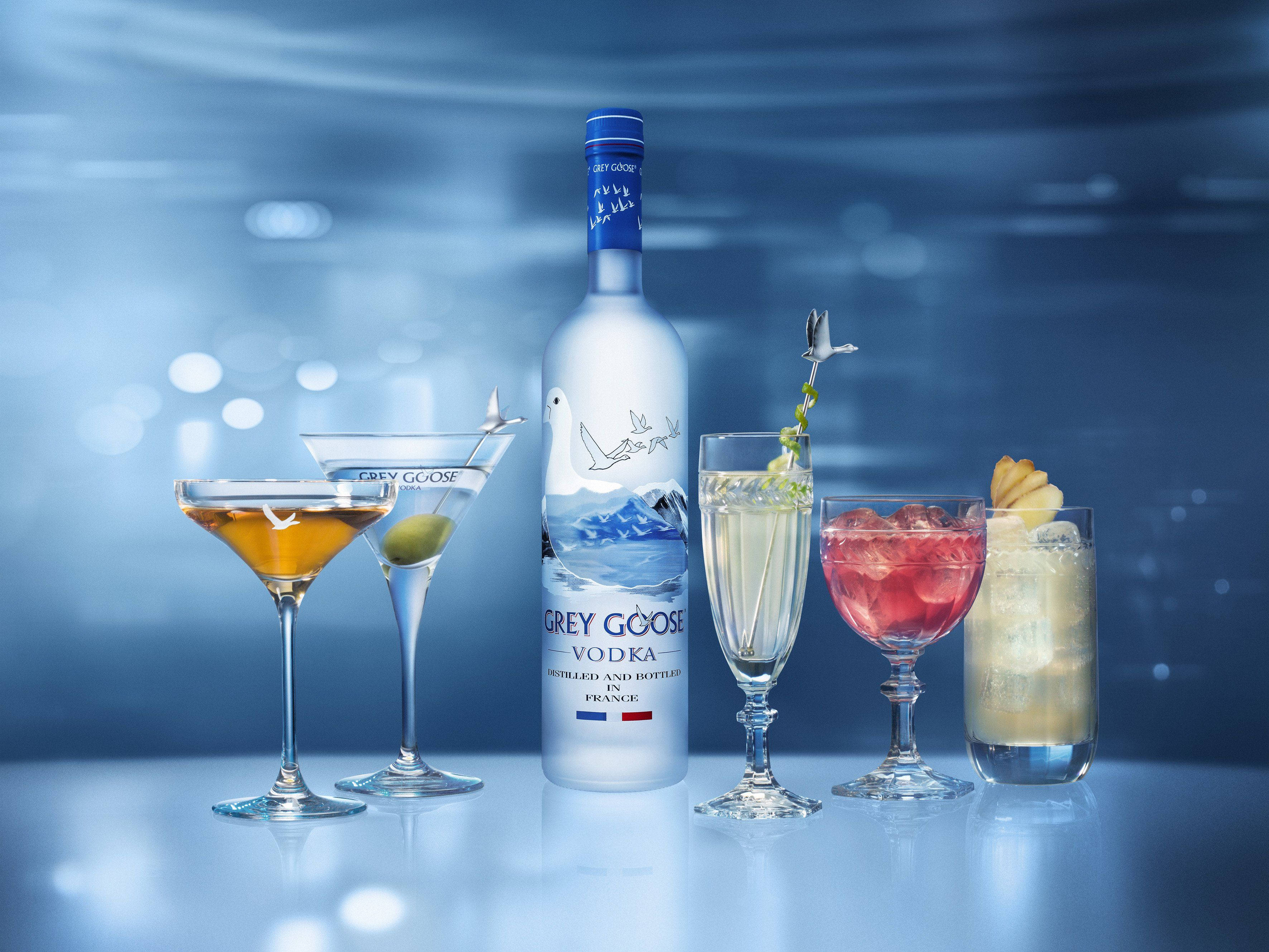 alcohol 桌布 壁紙 — 一個時尚的Grey Goose伏特加酒瓶，置於五杯優雅裝飾的雞尾酒中央，放在反射表面上，在冷藍色燈光下。