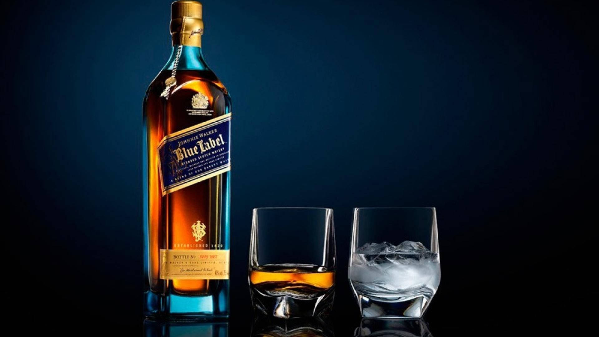  壁紙 — 一個時尚的Johnnie Walker Blue Label威士忌酒瓶旁邊有兩個玻璃杯，一個裝滿琥珀色液體，另一個裝有冰塊，背景是深藍色漸變。
