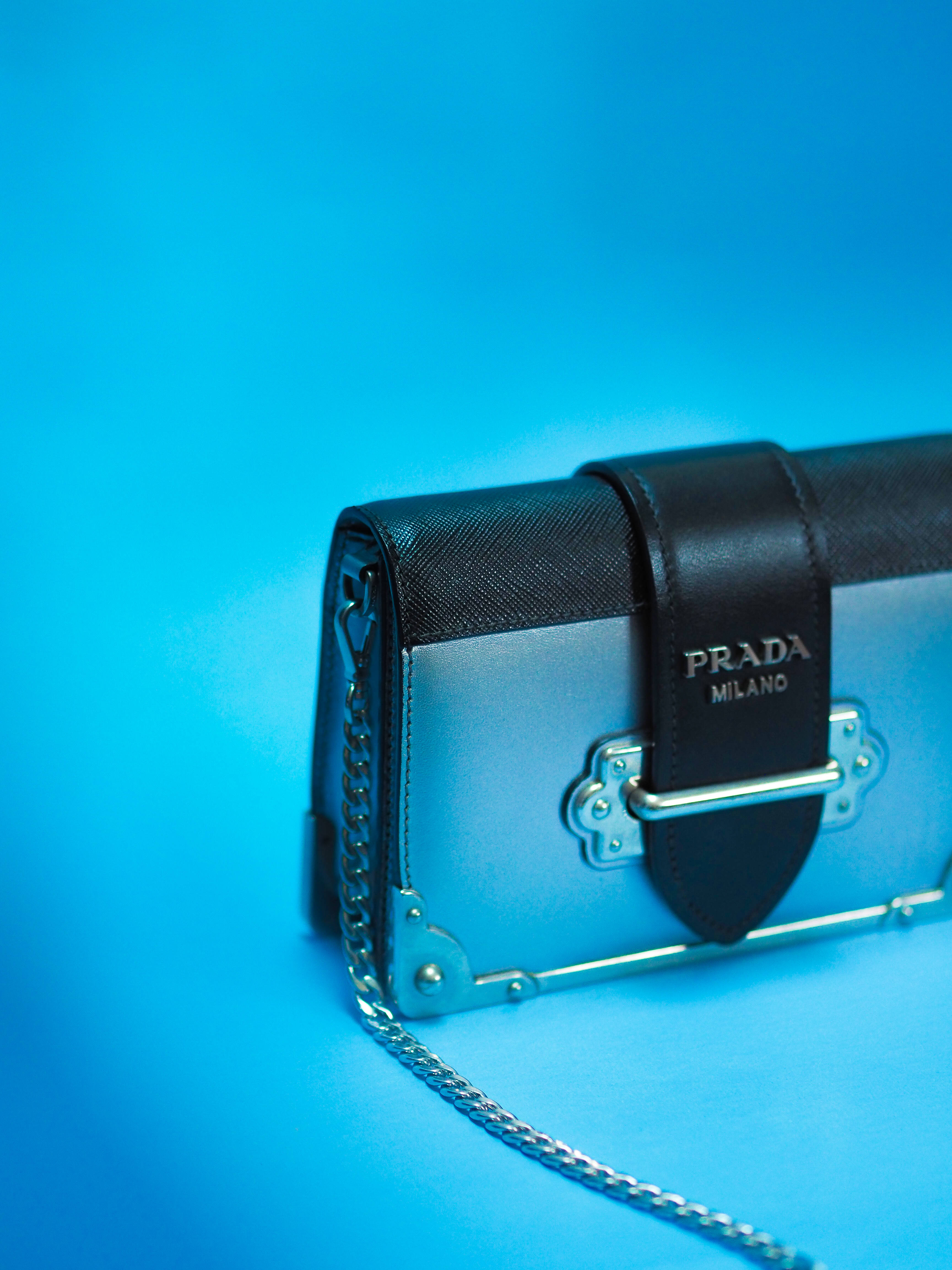 Prada 桌布 壁紙 — 一個時尚的Prada手提包，黑色皮革和銀色金屬配件，靠在充滿活力的藍色背景上。