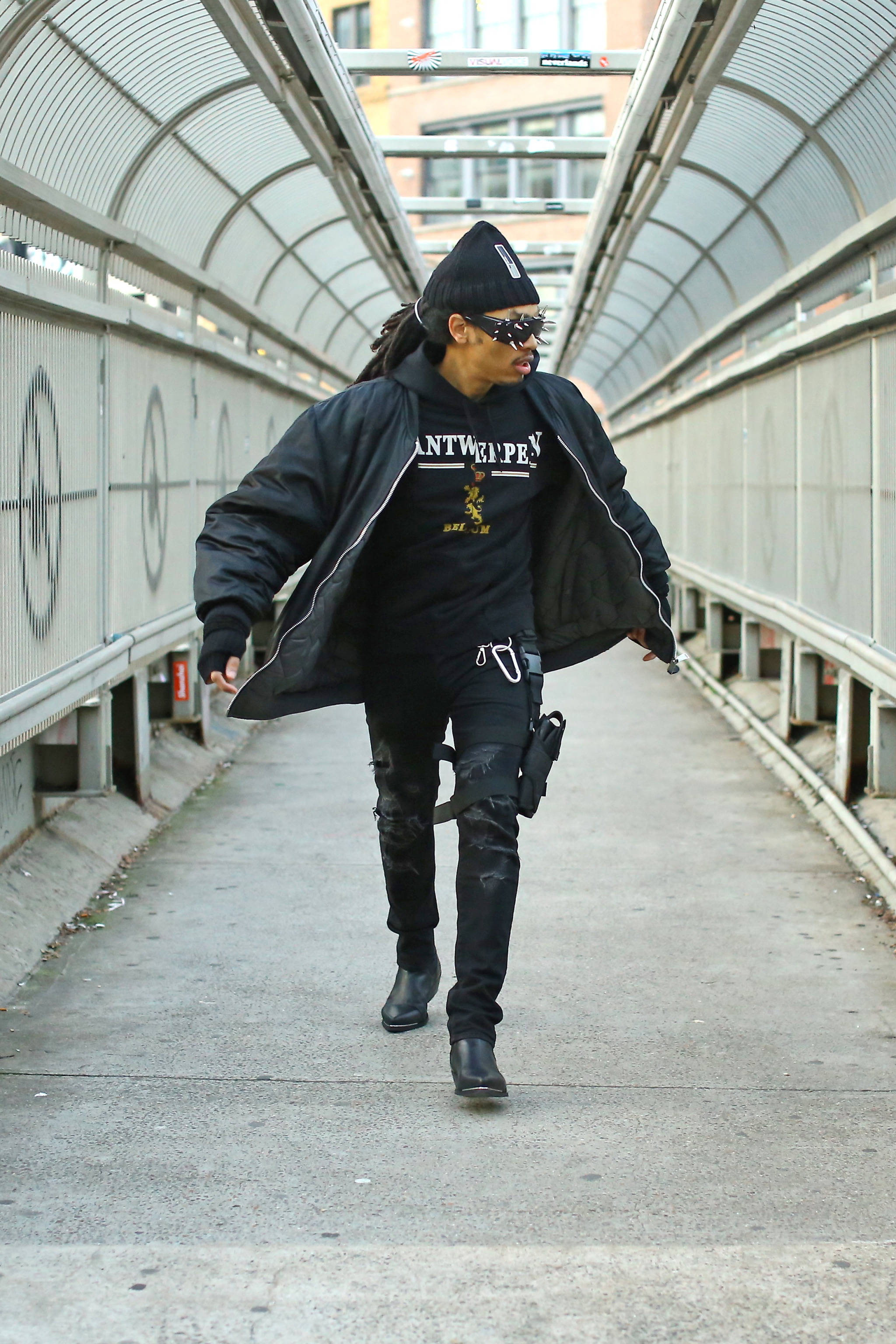 Vetements 桌布 壁紙 — 一個時尚的人穿著全黑，自信地走在現代有蓋行人天橋上。