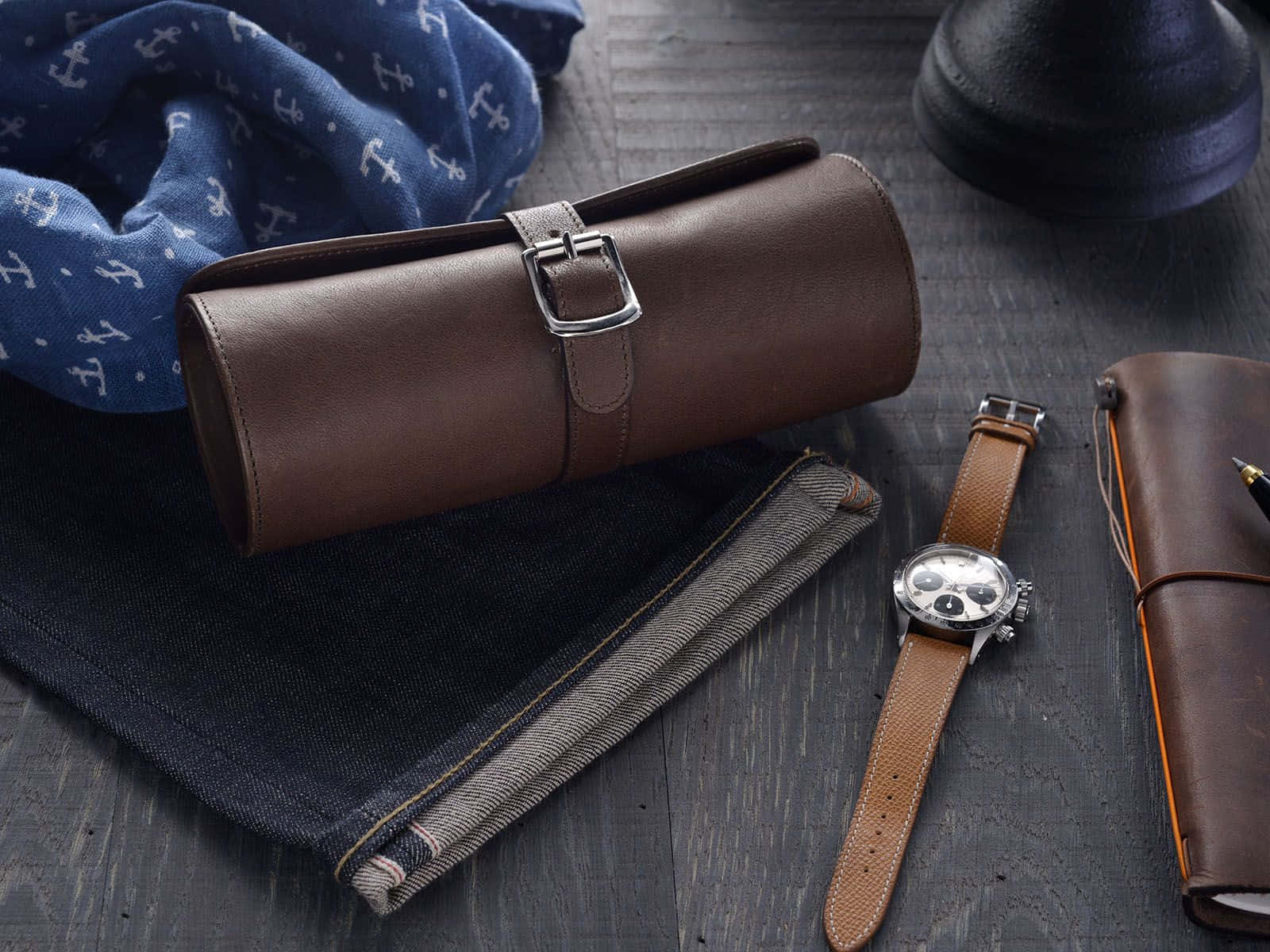 Accessory 桌布 壁紙 — 一個時尚的平鋪擺拍，包含棕色皮革手錶盒、計時碼錶、牛仔褲和藍色船錨圍巾，放置在深色木頭上。