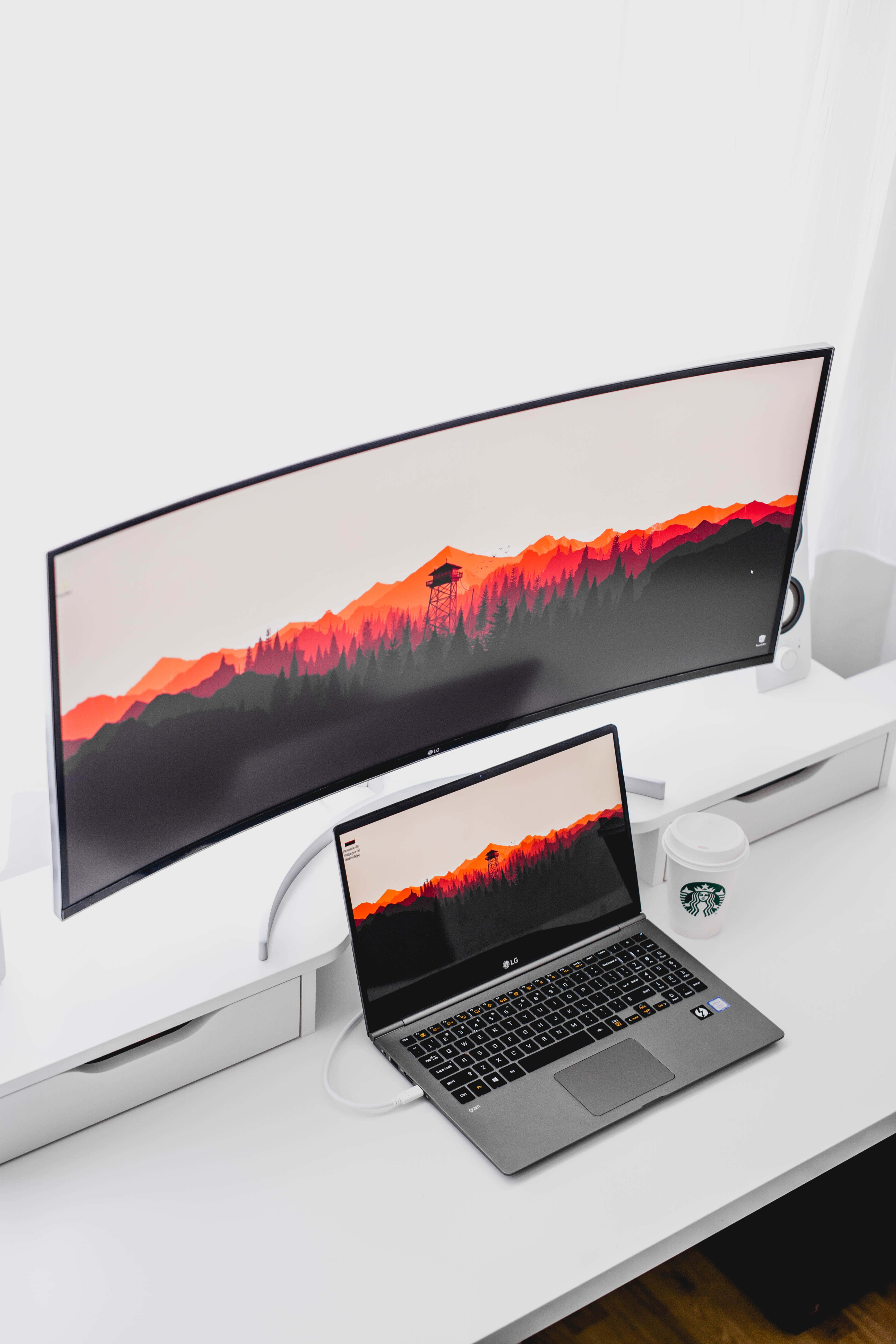 Monitor 桌布 壁紙 — 一個時尚的曲面顯示器和筆記型電腦在簡約的白色桌子上顯示充滿活力的橙色山景日落桌布。