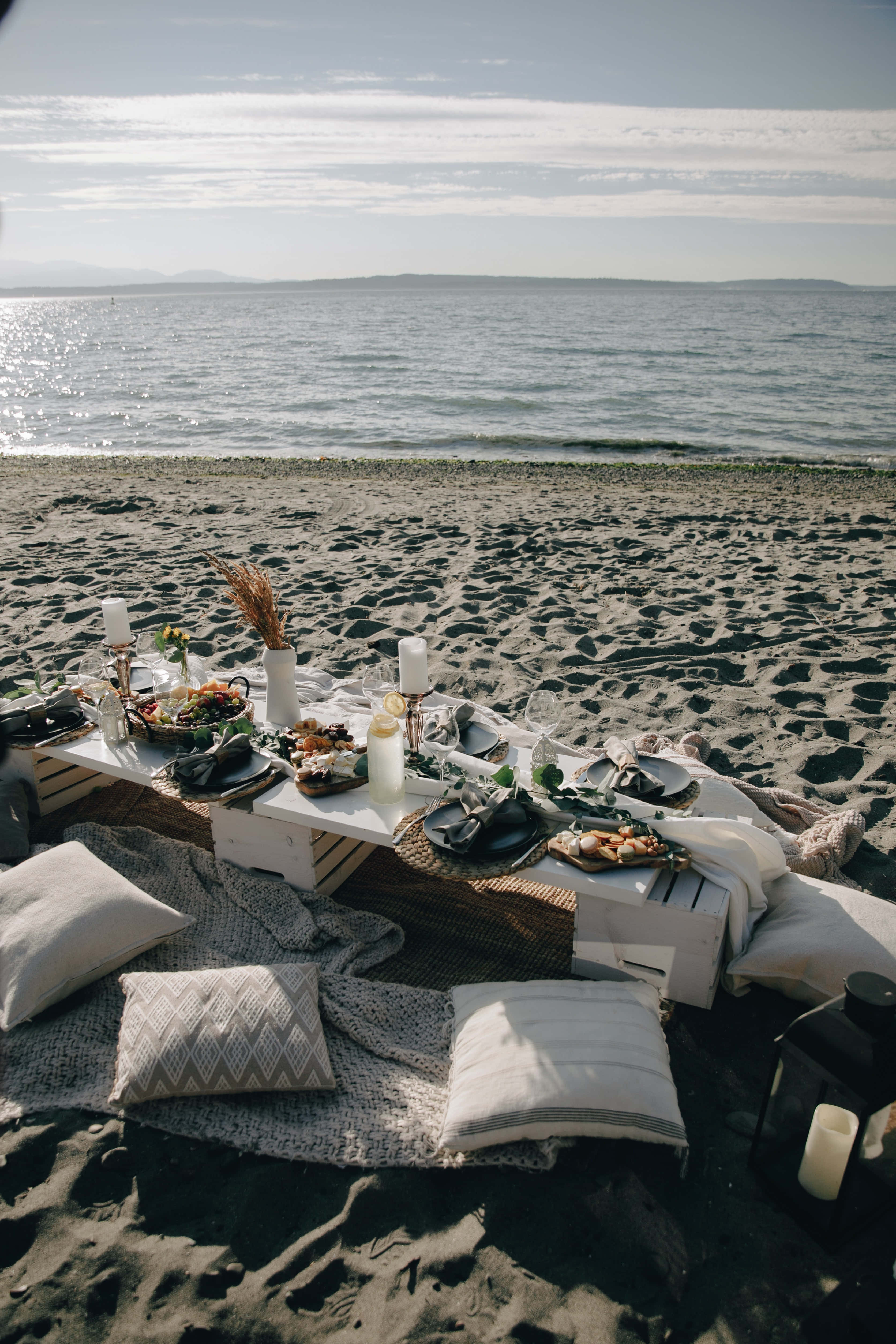Beach Picnic 桌布 壁紙 — 一個時尚的海灘野餐佈置，白色桌子上擺滿食物和蠟燭，置於沙灘上，在柔和日光下。