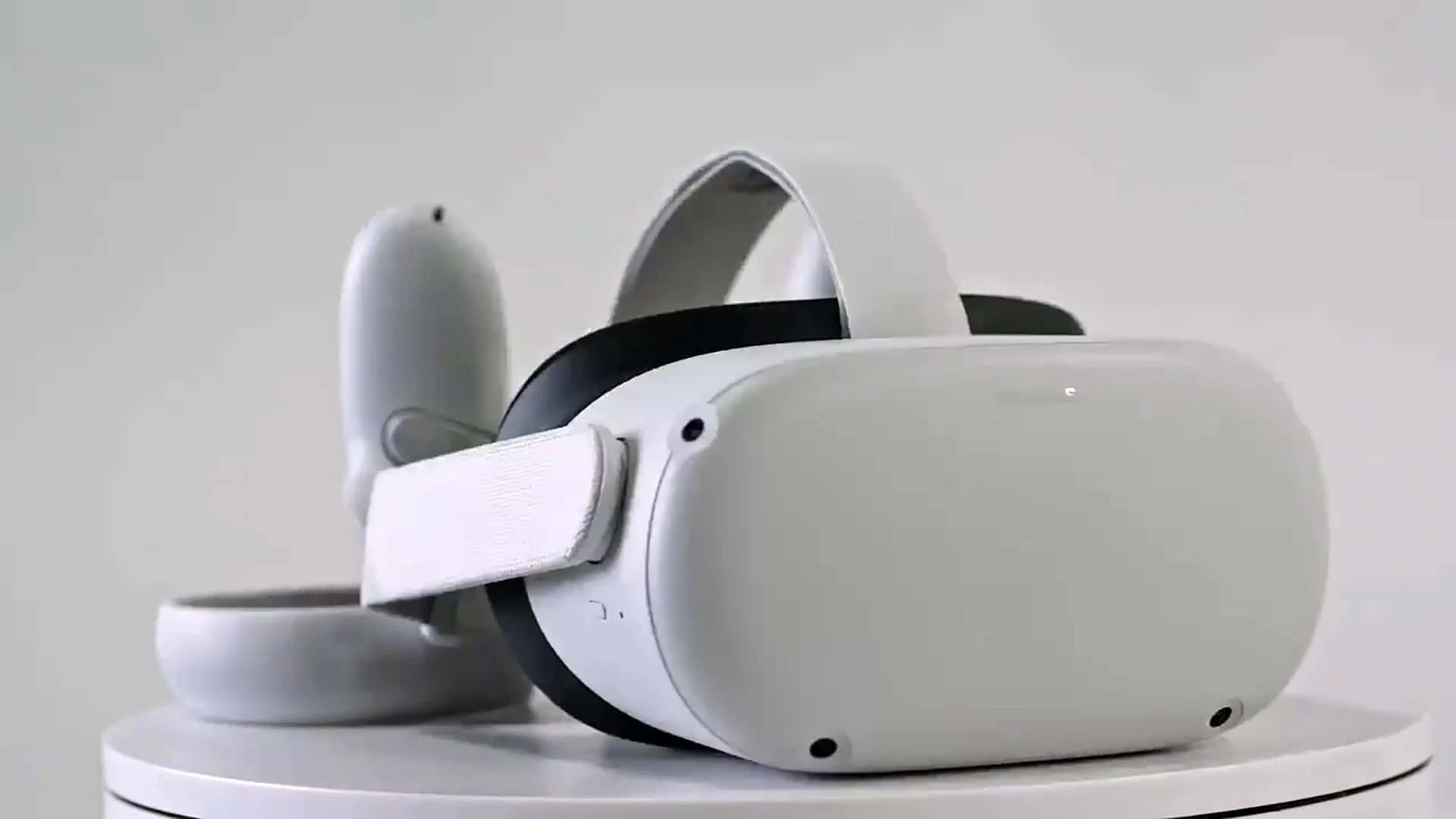 Oculus Quest 2 桌布 壁紙 — 一個時尚的白色VR頭戴式裝置連控制器，放置在簡約的白色底座上，背景乾淨。