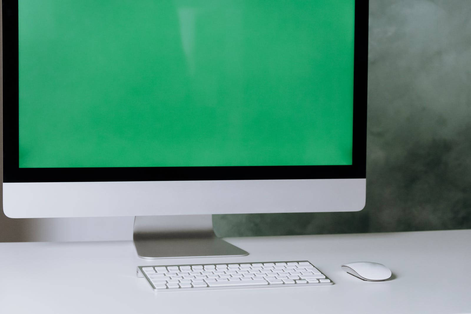 Green Screen 桌布 壁紙 — 一個時尚的白色電腦設置，配備綠色螢幕鍵盤和滑鼠，放在簡約的桌子上，背景是帶紋理的灰色牆壁。