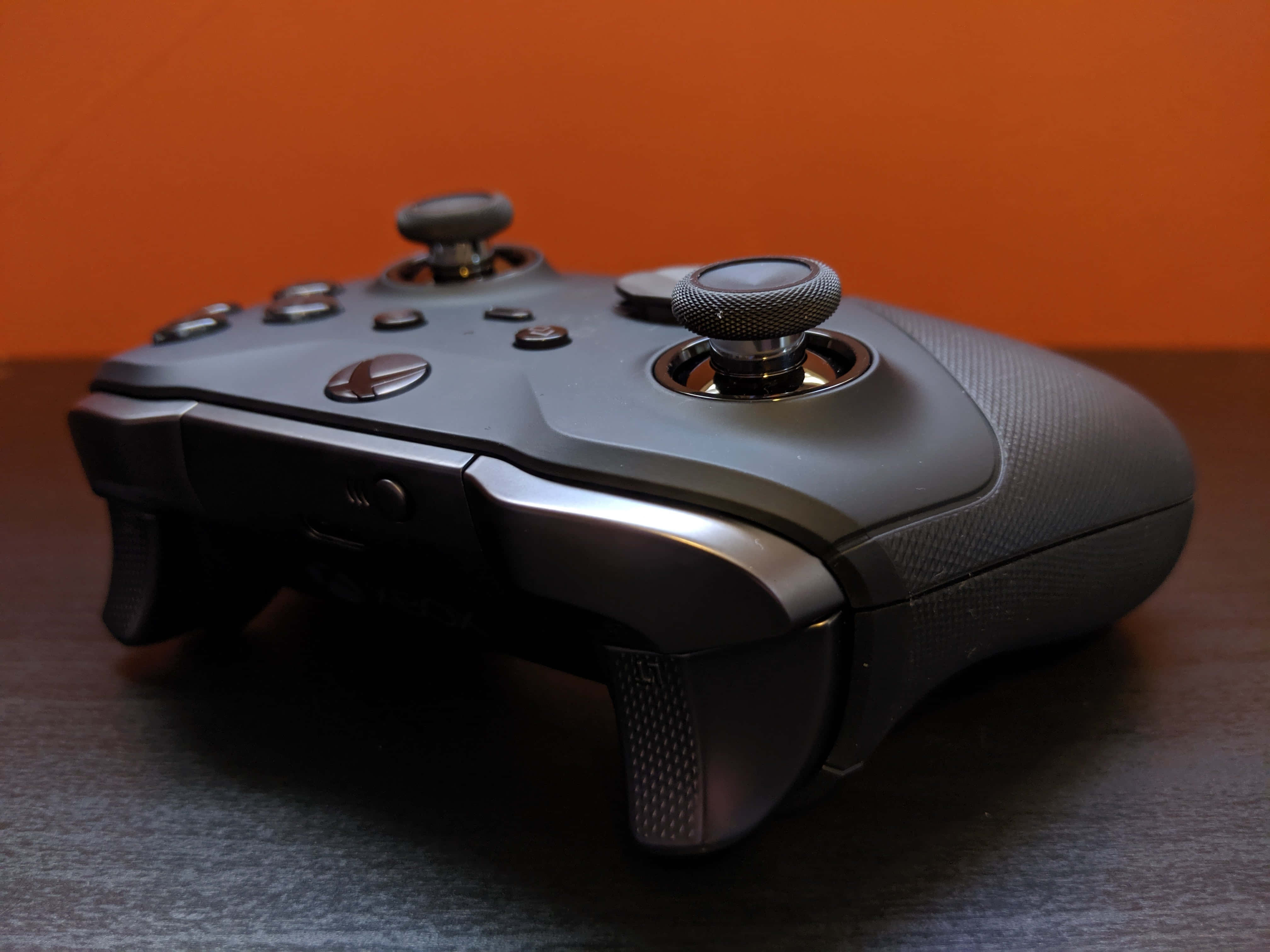Xbox 桌布 壁紙 — 一個時尚的磨砂黑色Xbox手把放在深色木質表面上，背景是溫暖的橙色。