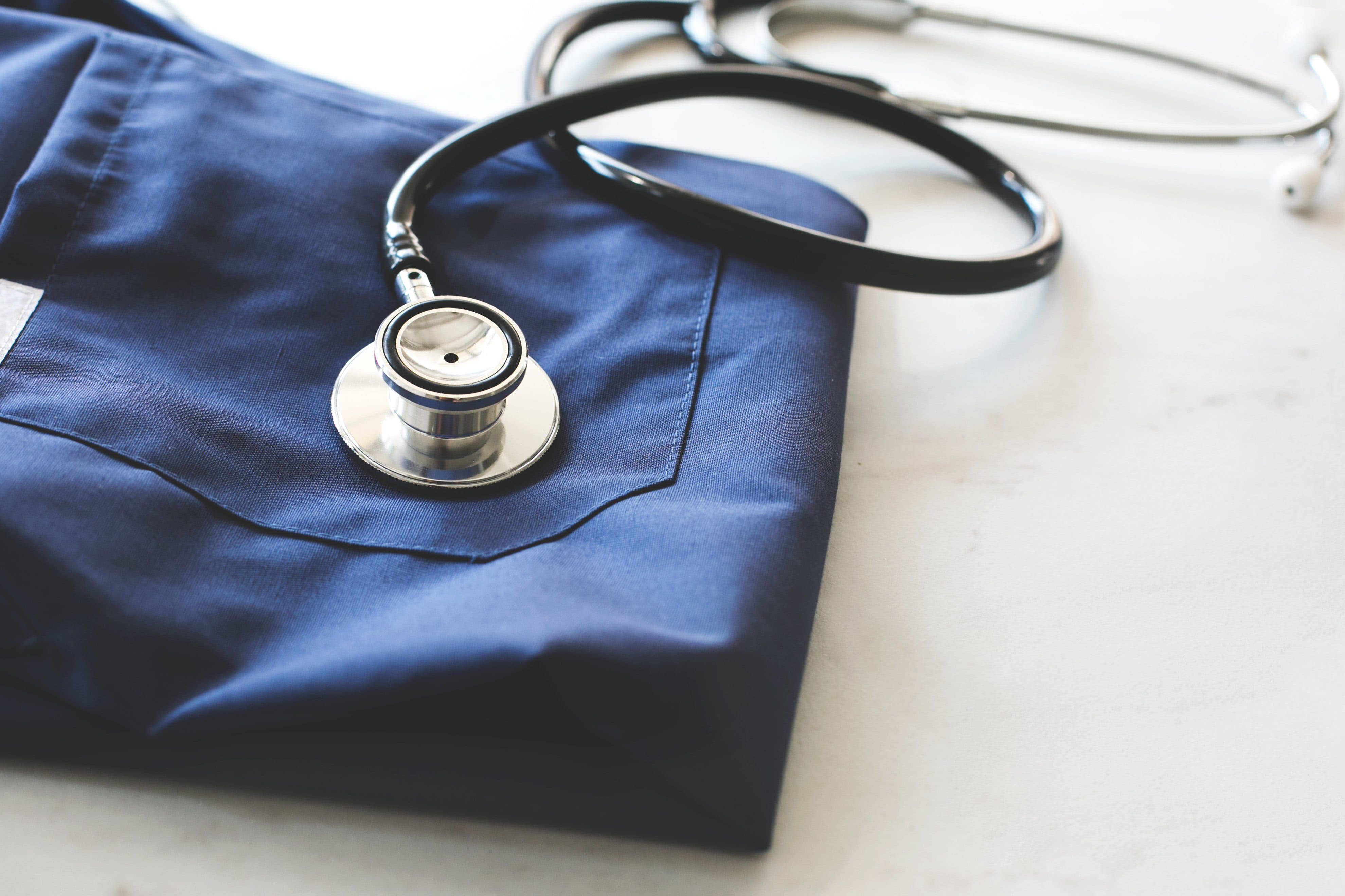 Stethoscope 桌布 壁紙 — 一個時尚的聽診器放在折疊好的海軍藍色醫用刷手服上，置於潔白的表面上。