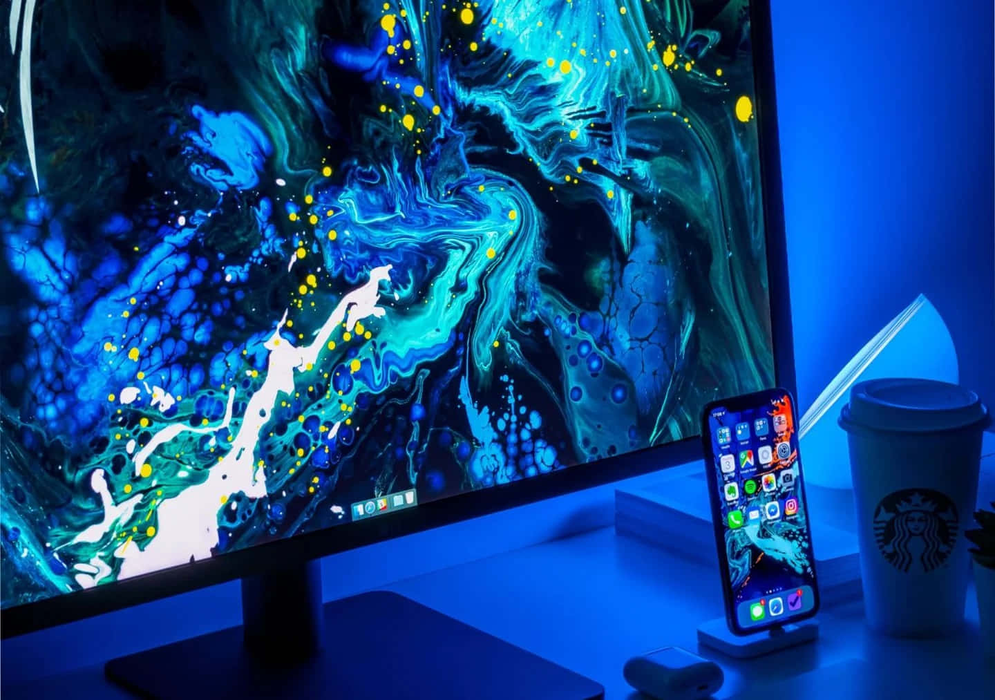 Computer Monitor 桌布 壁紙 — 一個時尚的辦公桌設置沐浴在藍光中，有一個大顯示器顯示充滿活力的抽象藍色和黃色流體藝術壁紙，以及一部智能手機和星巴克杯子。