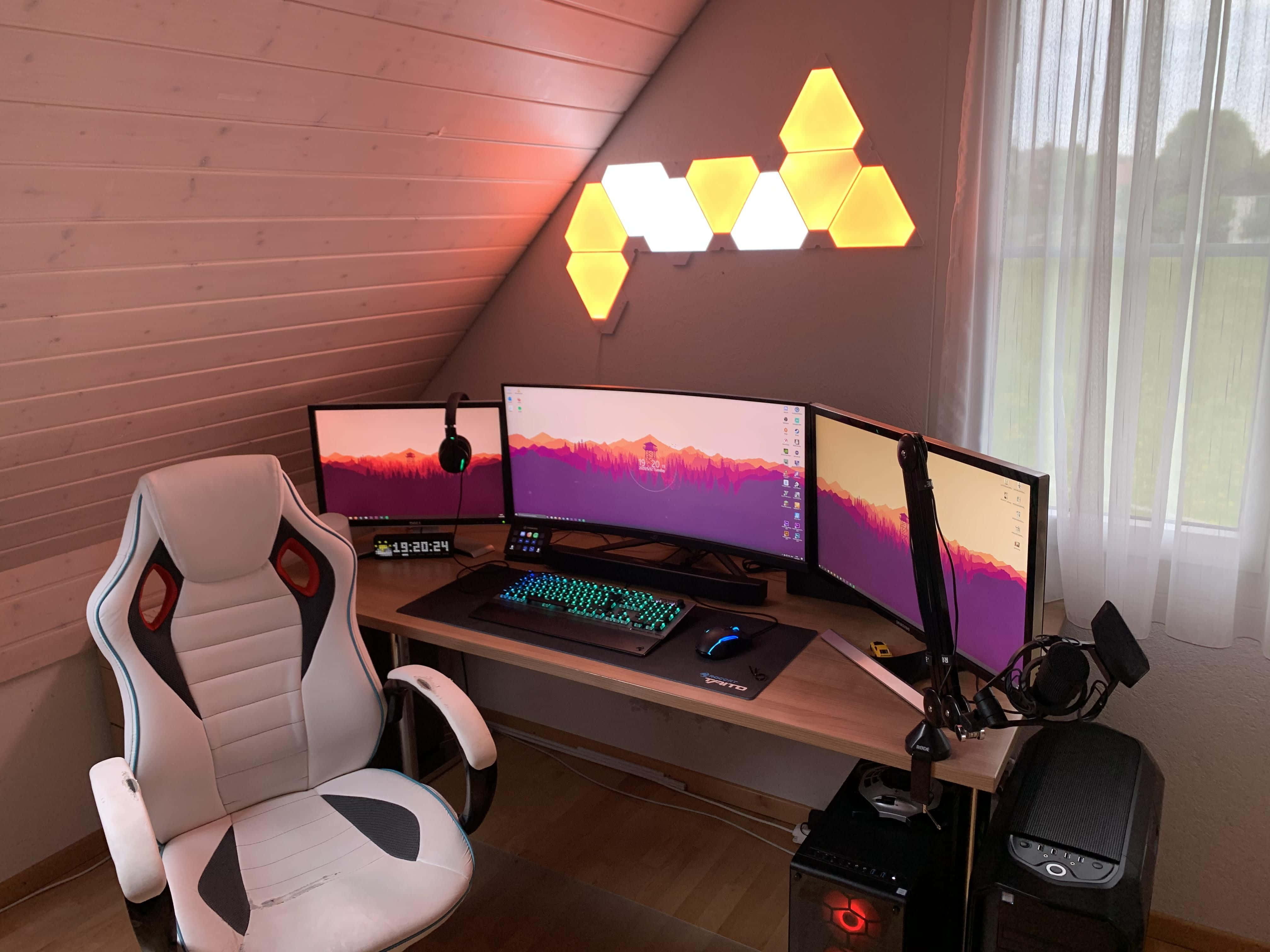 Gaming Pc Setup 桌布 壁紙 — 一個時尚的遊戲設置，配有三個顯示器，顯示山景日落，由幾何牆燈和發光鍵盤照亮。