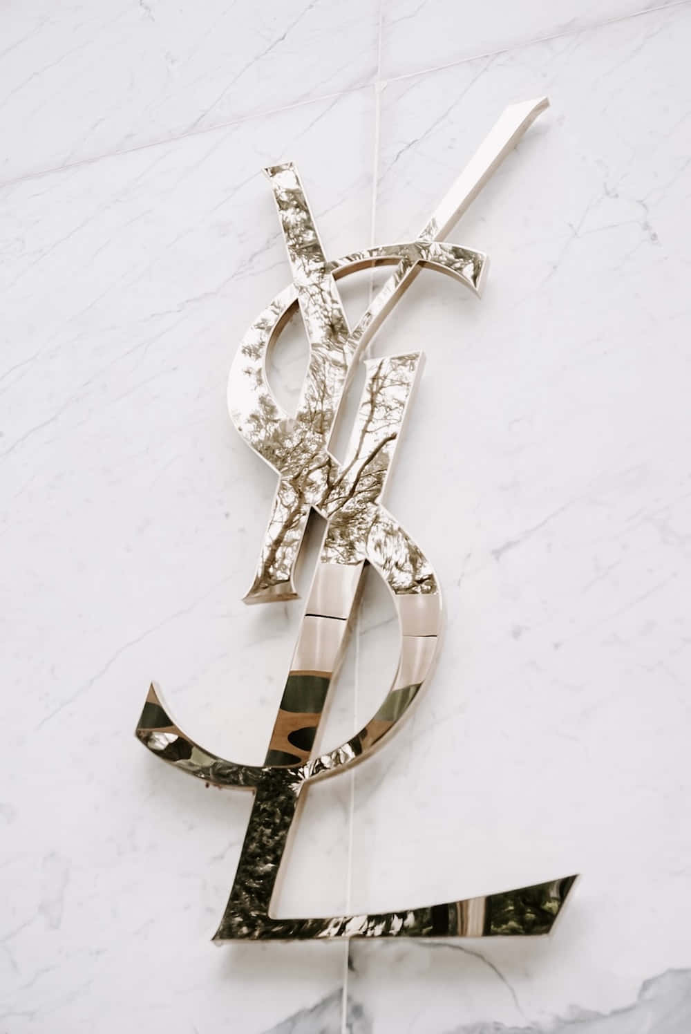 YSL 桌布 壁紙 — 一個時尚的金色YSL標誌雕塑，帶有紋理金屬飾面，安裝在白色大理石牆上。