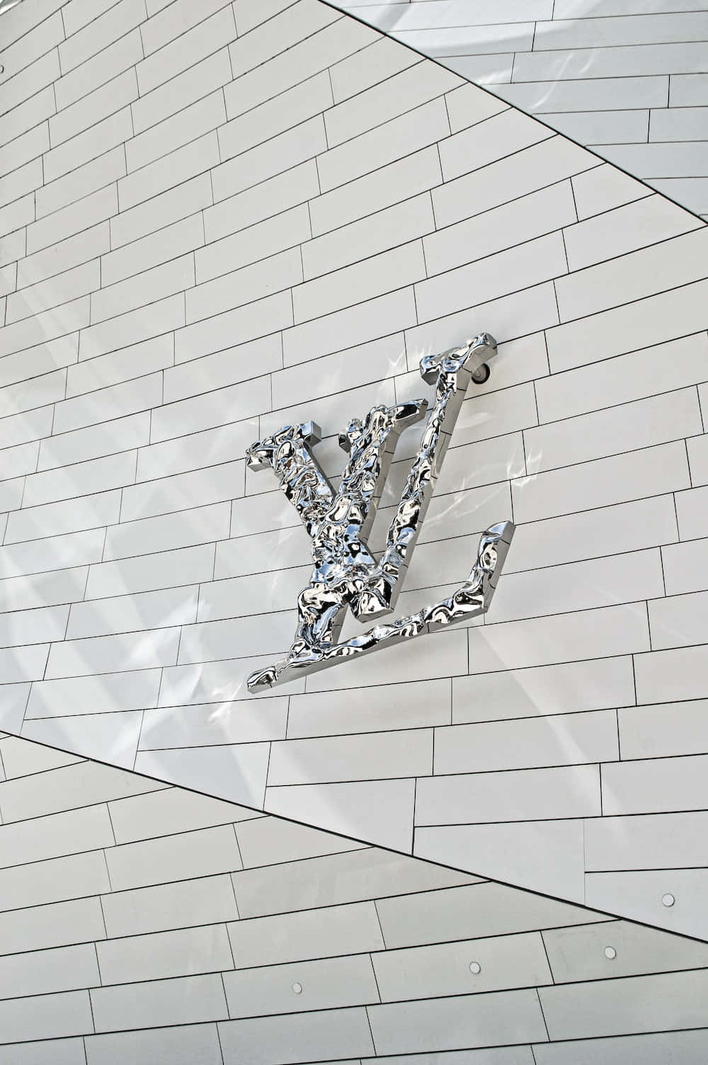 路易威登 桌布 壁紙 — 一個時尚的鉻色 Louis Vuitton 標誌安裝在白色瓷磚牆上，帶有幾何角度和銳利陰影。