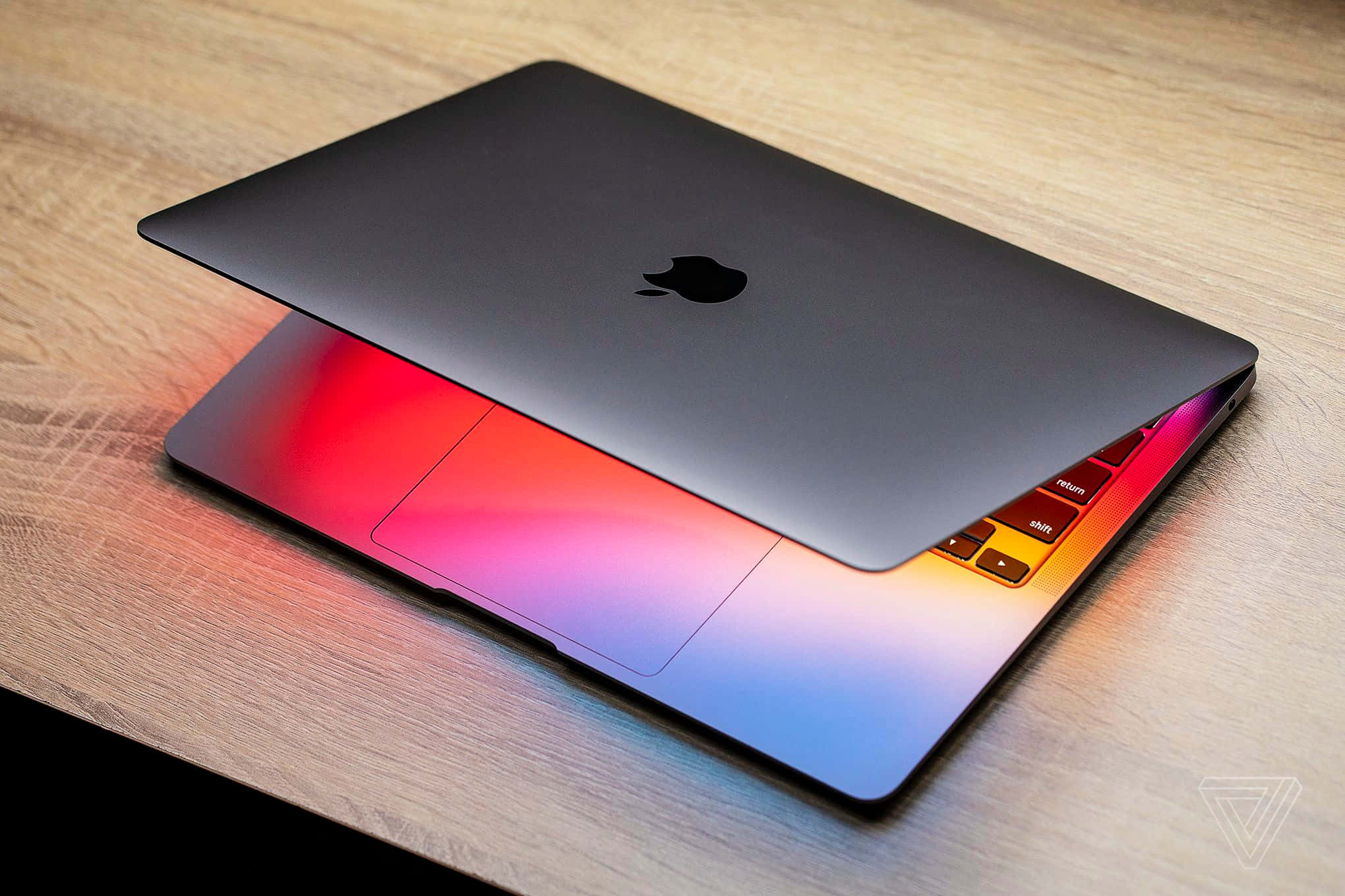 MacBook 動態圖片 桌布 壁紙 — 一個時尚的銀色MacBook蓋子微微打開，放在木桌上，露出鍵盤背光燈發出的鮮豔彩虹光芒。