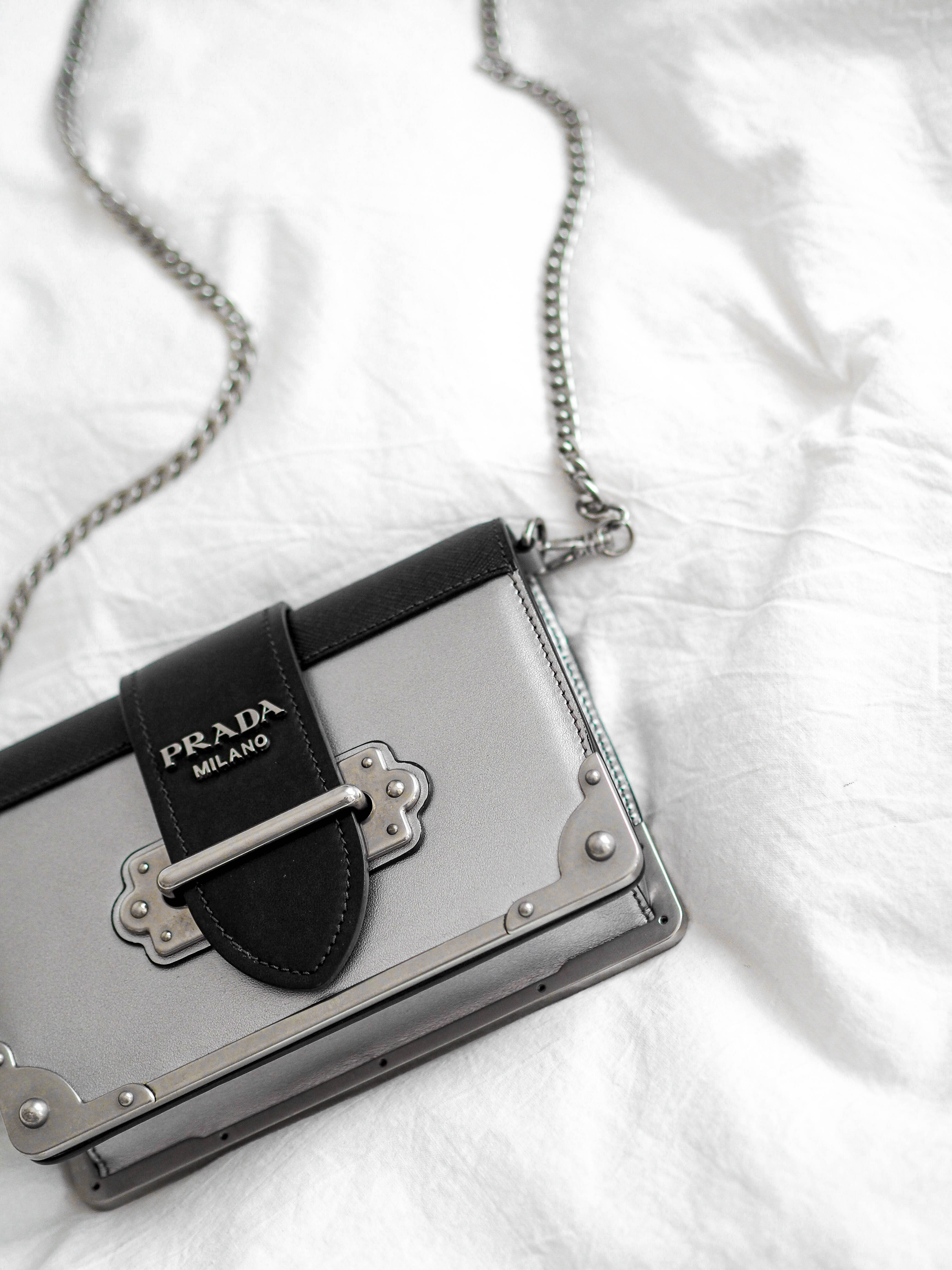 Prada 桌布 壁紙 — 一個時尚的銀色Prada手提包，配有黑色皮革飾邊和鏈條肩帶，放在皺巴巴的白色布料上。