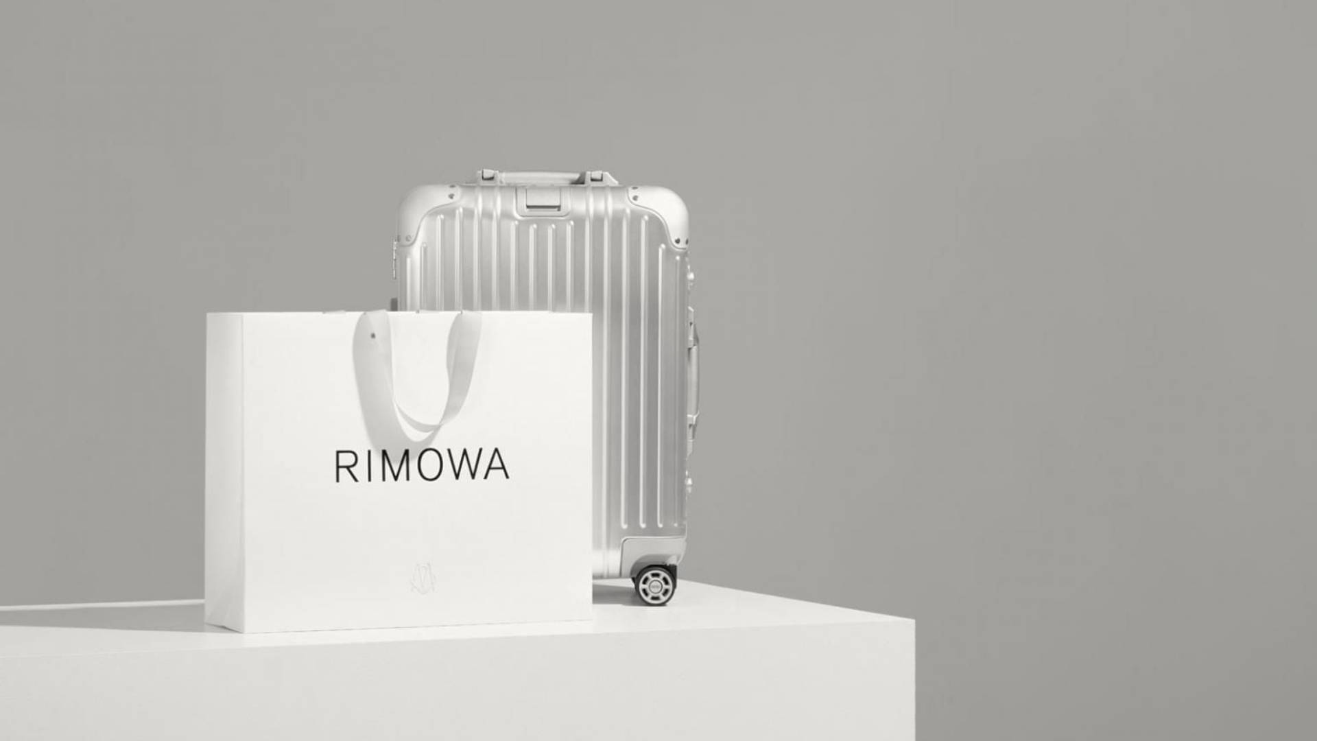 品牌 桌布 壁紙 — 一個時尚的銀色Rimowa行李箱與其品牌白色紙袋一起放在簡約的白色底座上，背景為中性灰色。