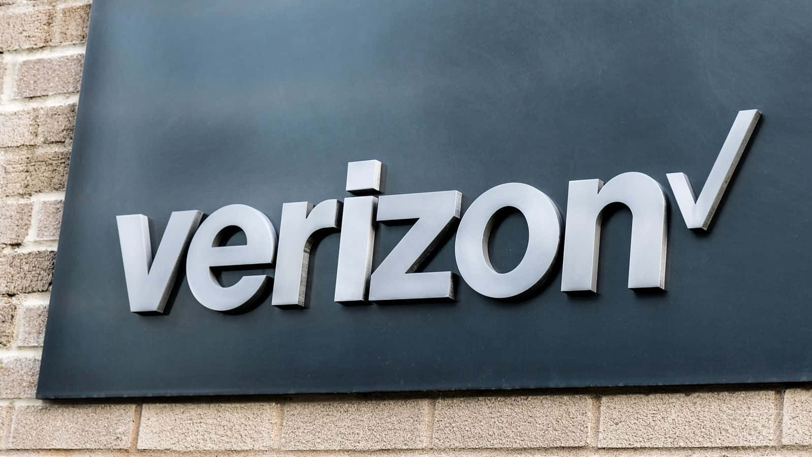 威訊 桌布 壁紙 — 一個時尚的銀色Verizon標誌安裝在深色面板上，背景是淺色磚牆。