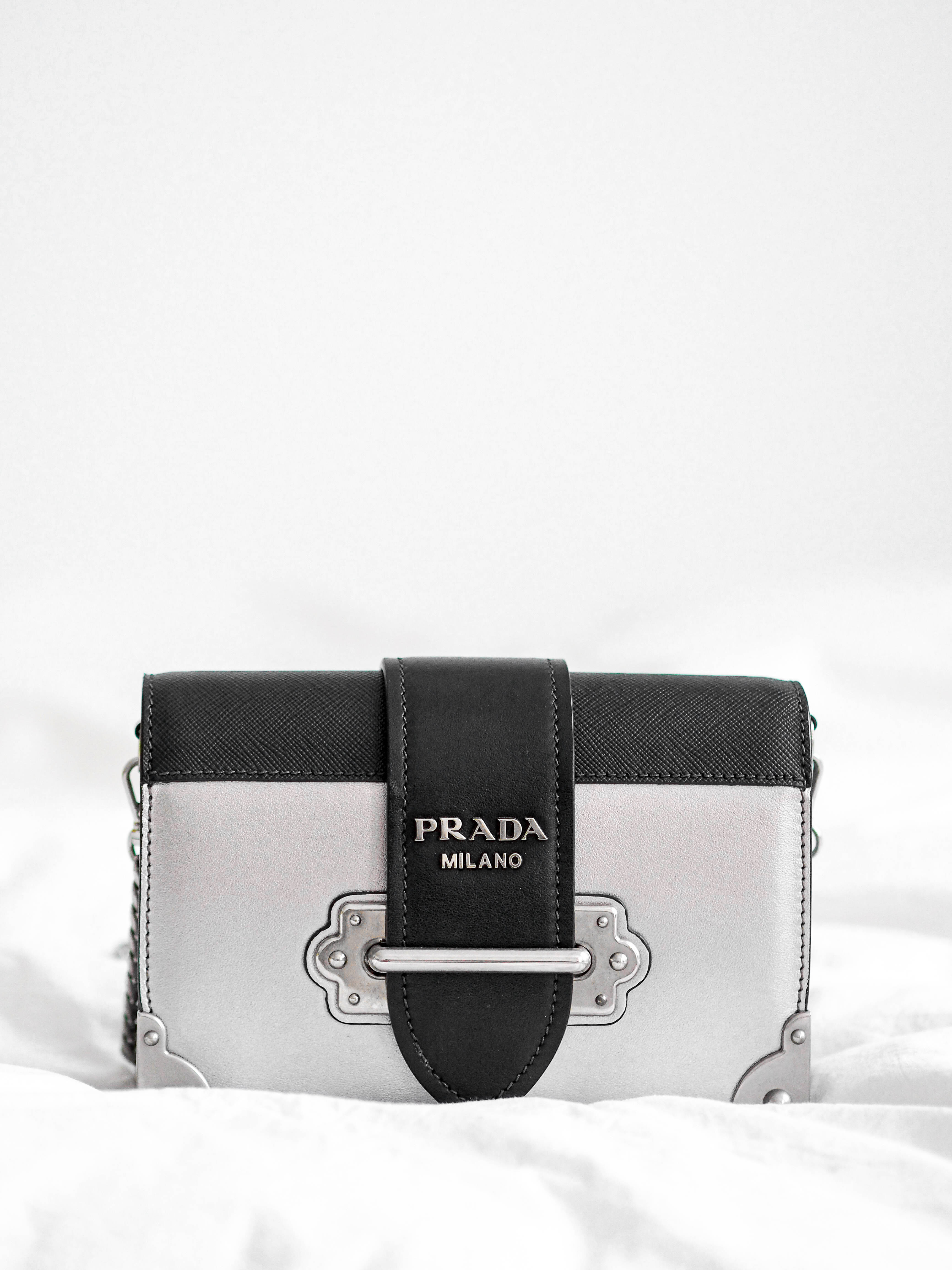 Prada 桌布 壁紙 — 一個時尚的銀色和黑色Prada手提包，帶有金屬配件，放在柔軟的白色布料背景上。