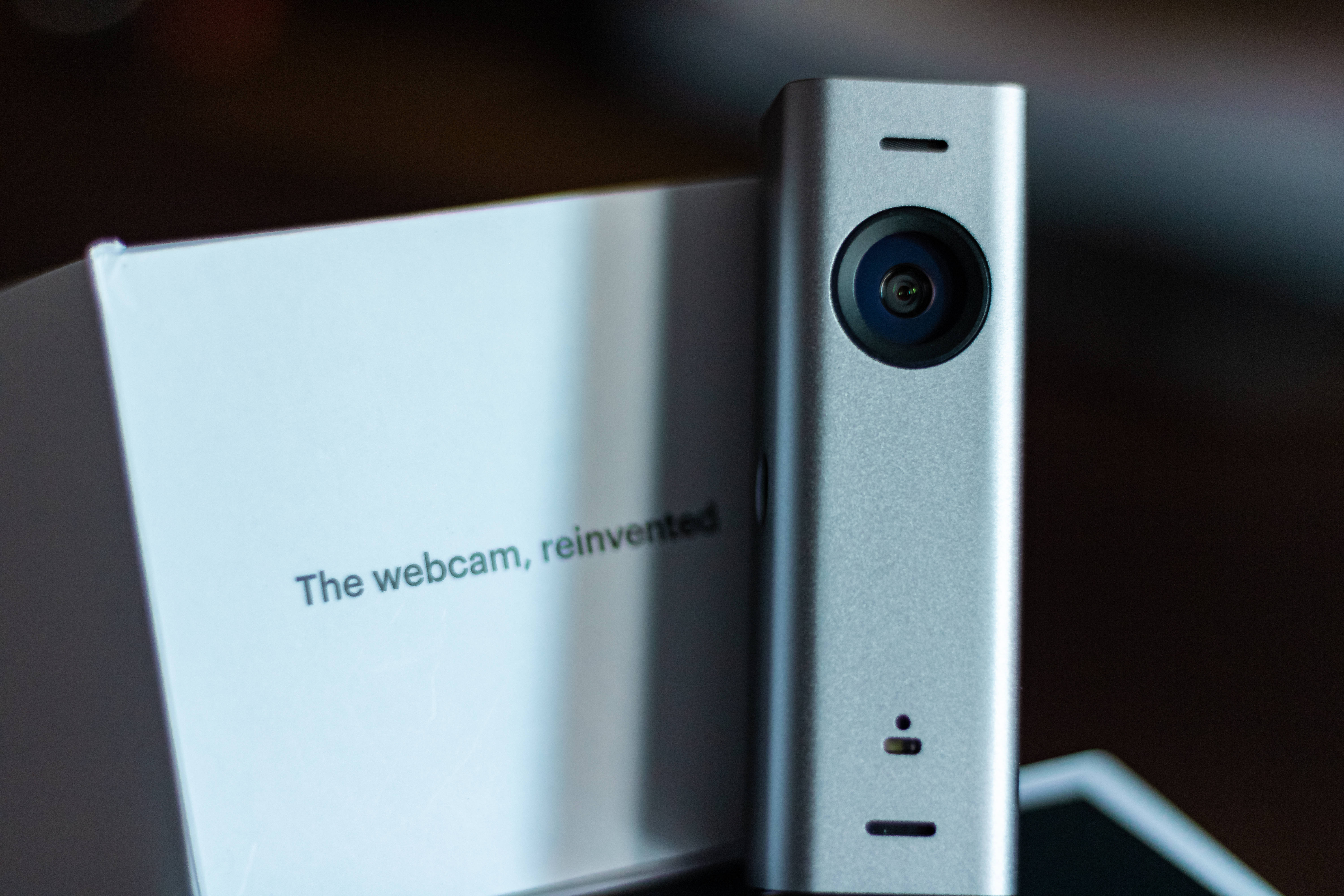 Webcam 桌布 壁紙 — 一個時尚的銀色網路攝影機站在其白色盒子旁邊，盒子上標有 'The webcam reinvented'，在柔和的攝影棚燈光下。