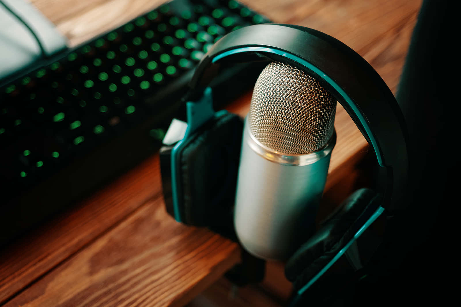 Podcast 桌布 壁紙 — 一個時尚的銀色麥克風和黑色遊戲耳機放在木桌上，旁邊是一個發光的綠色鍵盤。