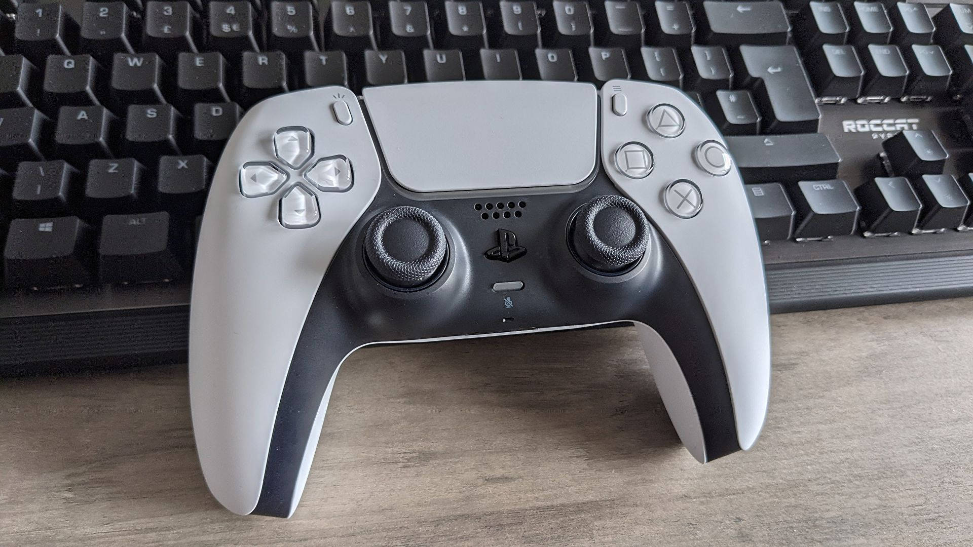 PS5 動態圖片 桌布 壁紙 — 一個時尚的銀黑色PlayStation 5 DualSense控制器放在木桌上，旁邊有一個黑色鍵盤。