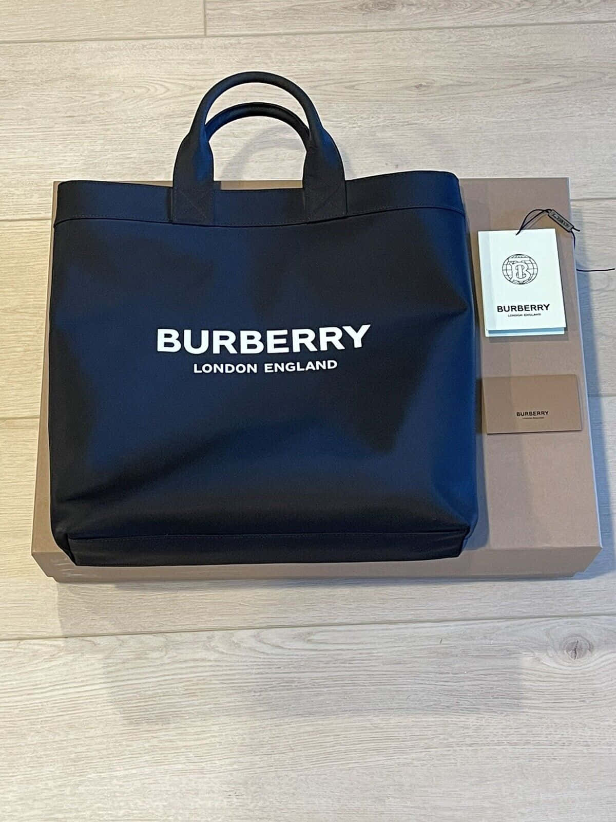 Burberry 桌布 壁紙 — 一個時尚的黑色 Burberry 手提袋，帶有白色標誌，放在其品牌盒子上，位於淺色木地板上。