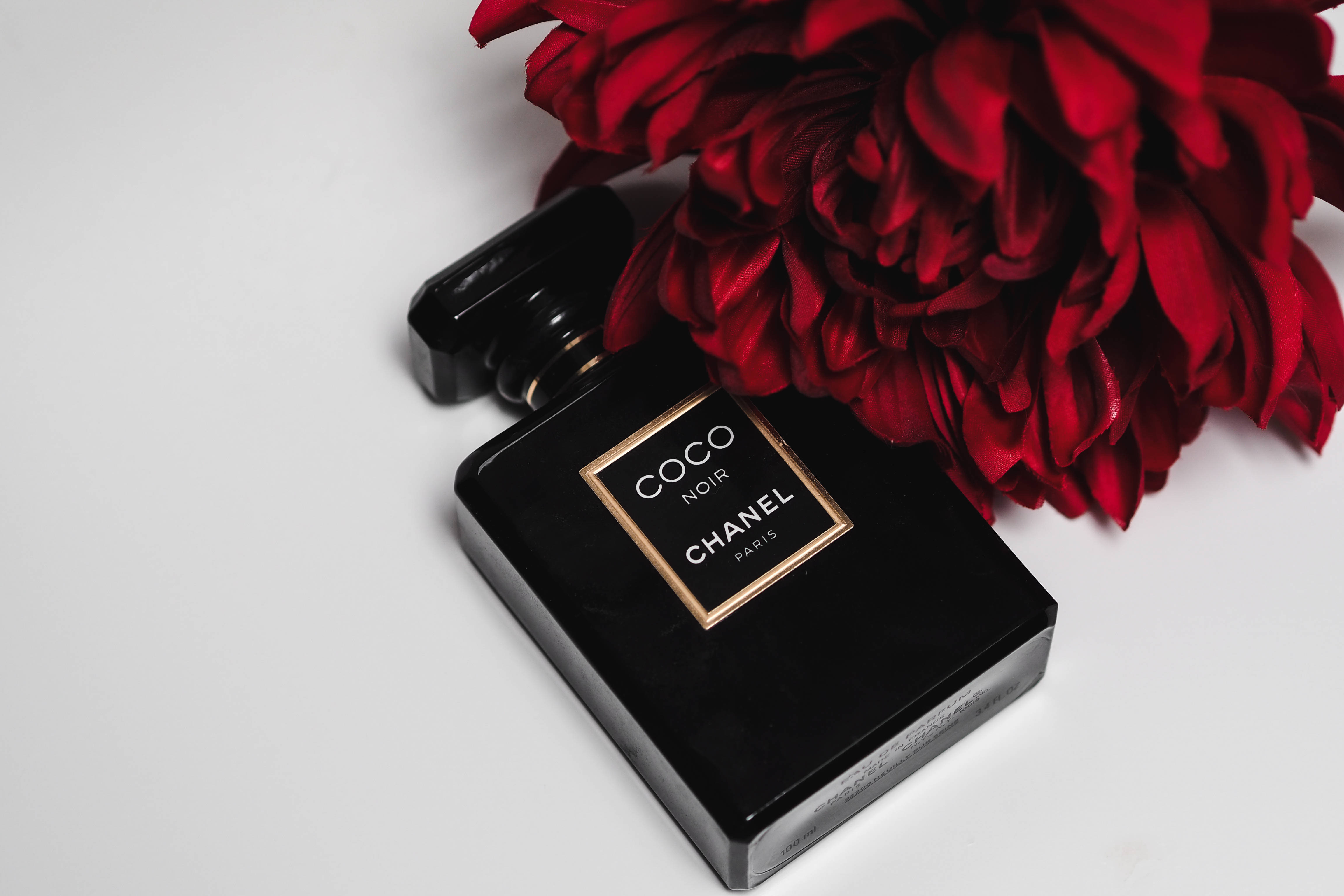 Chanel 桌布 壁紙 — 一個時尚的黑色 Chanel Coco Noir 香水瓶放在純白表面上，旁邊是一朵茂盛的紅花。