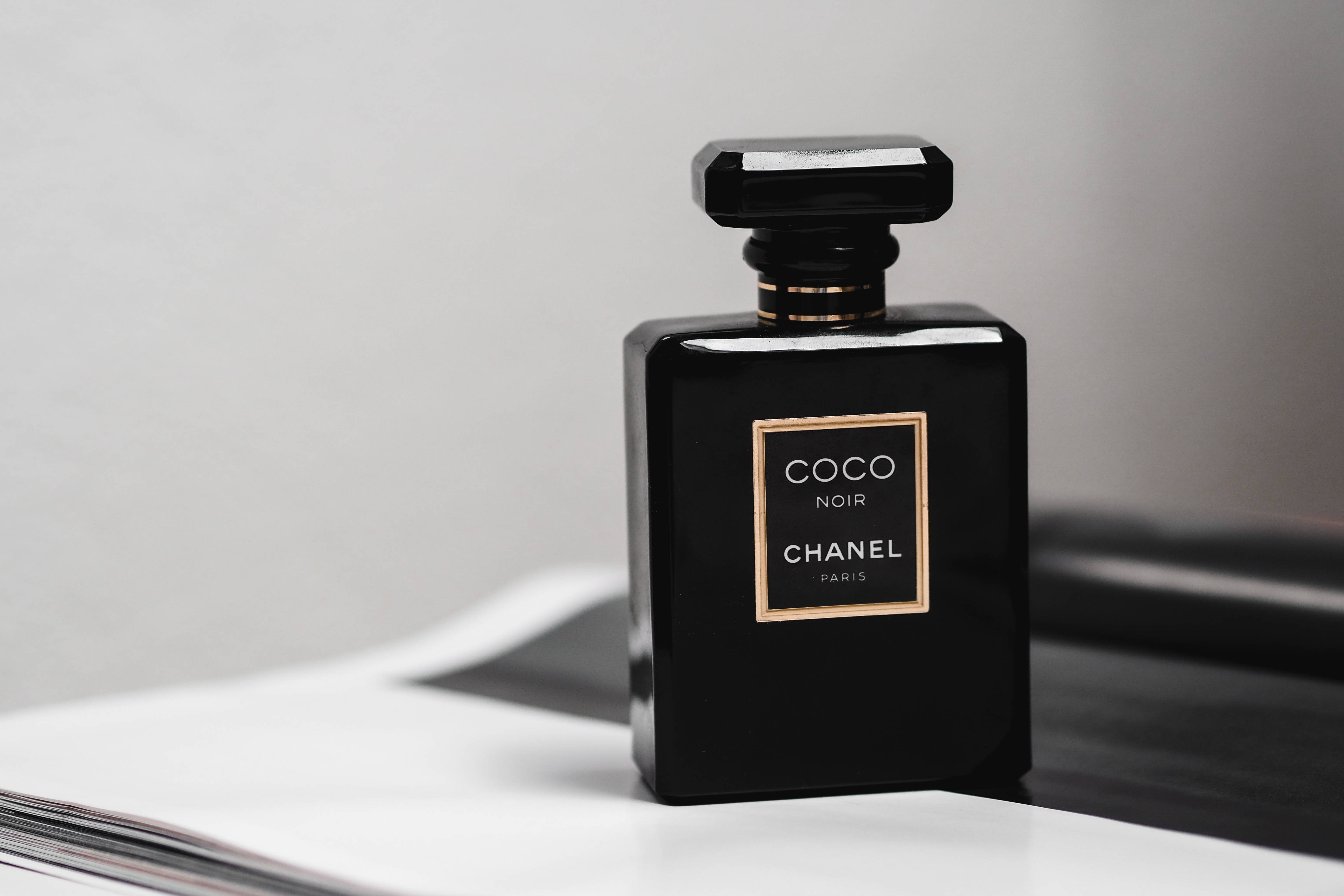 Chanel 桌布 壁紙 — 一個時尚的黑色 Chanel Coco Noir 香水瓶，帶有金色點綴，優雅地放置在白色表面上，背景為柔和的灰色。