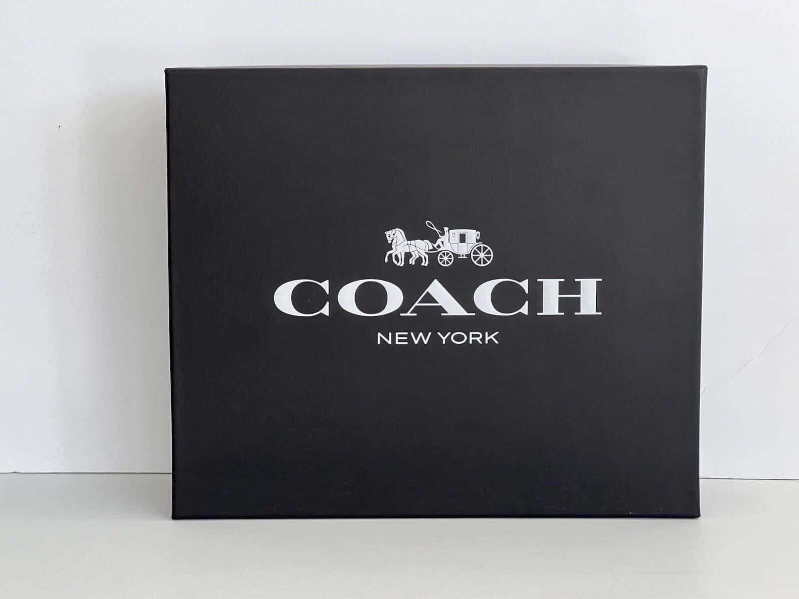 教練 桌布 壁紙 — 一個時尚的黑色 Coach New York 盒子，帶有白色標誌，居中放置在白色表面上，背景是純白色牆壁。