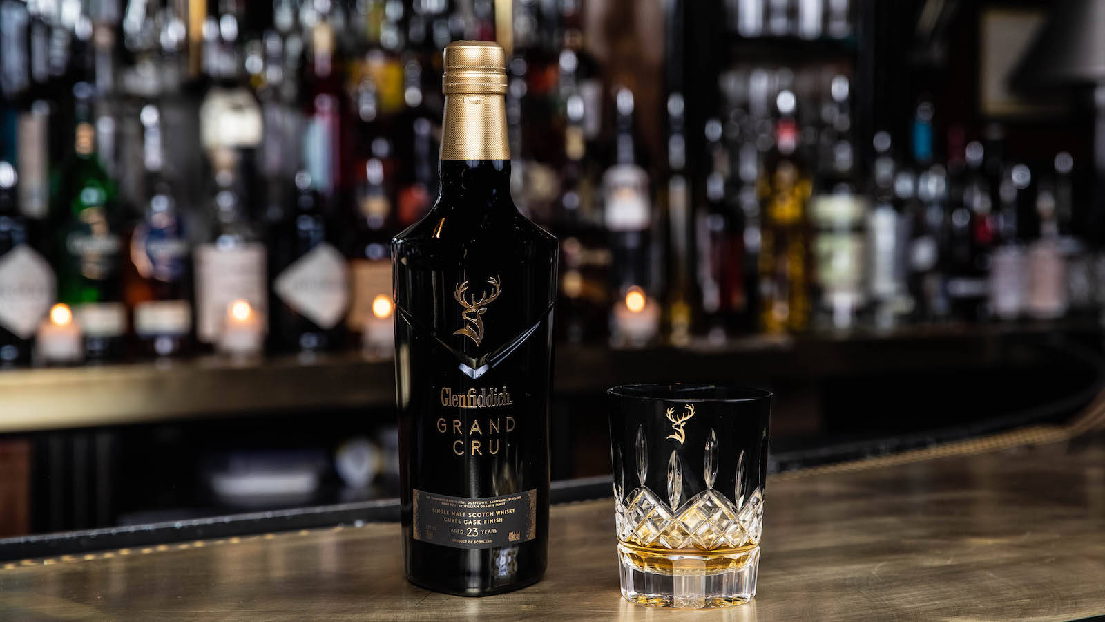 飲料 桌布 壁紙 — 一個時尚的黑色 Glenfiddich Grand Cru 威士忌酒瓶和一個裝滿琥珀色液體的水晶杯放在拋光酒吧檯上，背景有模糊的酒瓶和溫暖的燭光。