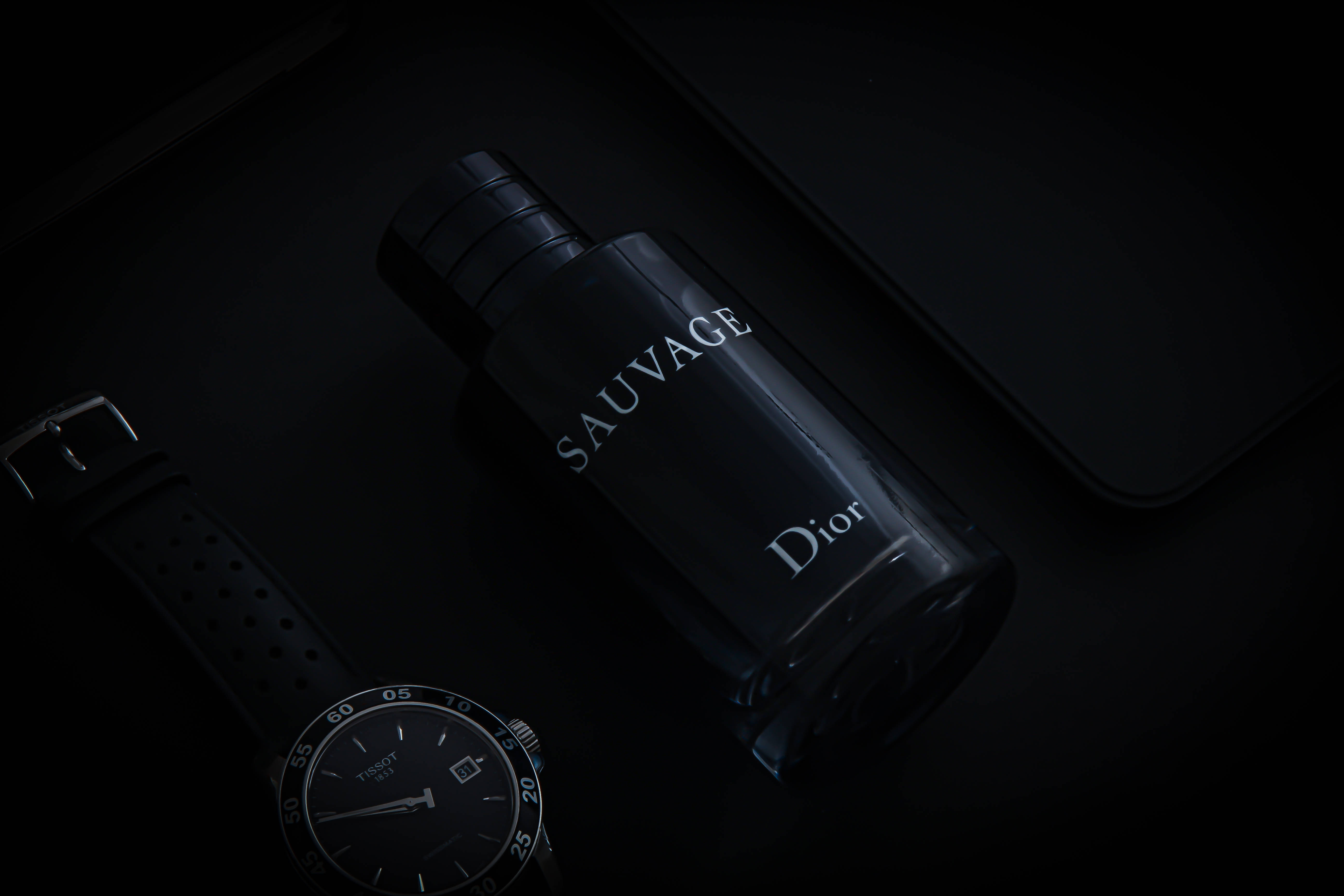 Dior 動態 桌布 壁紙 — 一個時尚的黑色Dior Sauvage香水瓶在戲劇性燈光下，放在深色表面上，旁邊有一隻相配的手錶。