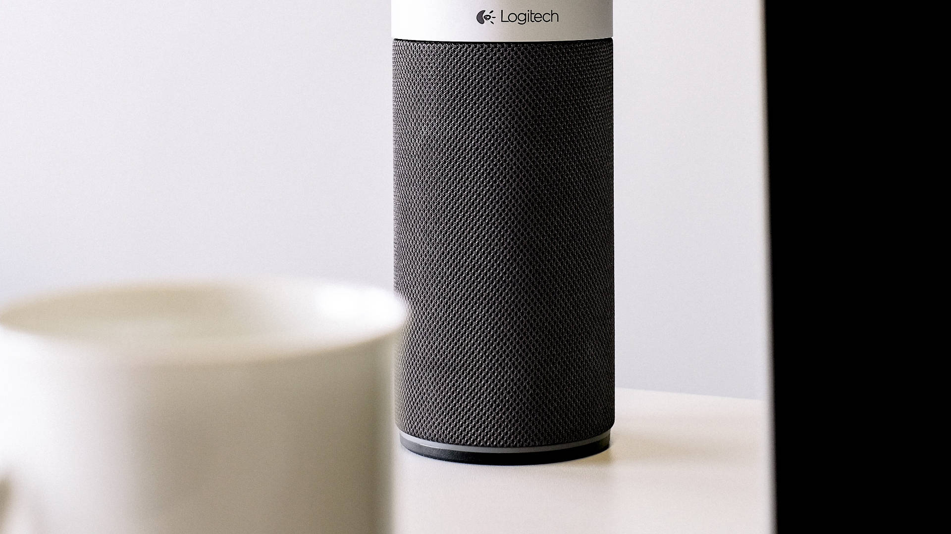 Logitech 桌布 壁紙 — 一個時尚的黑色Logitech揚聲器放在白色表面上，旁邊是一個模糊的白色杯子，背景簡約。
