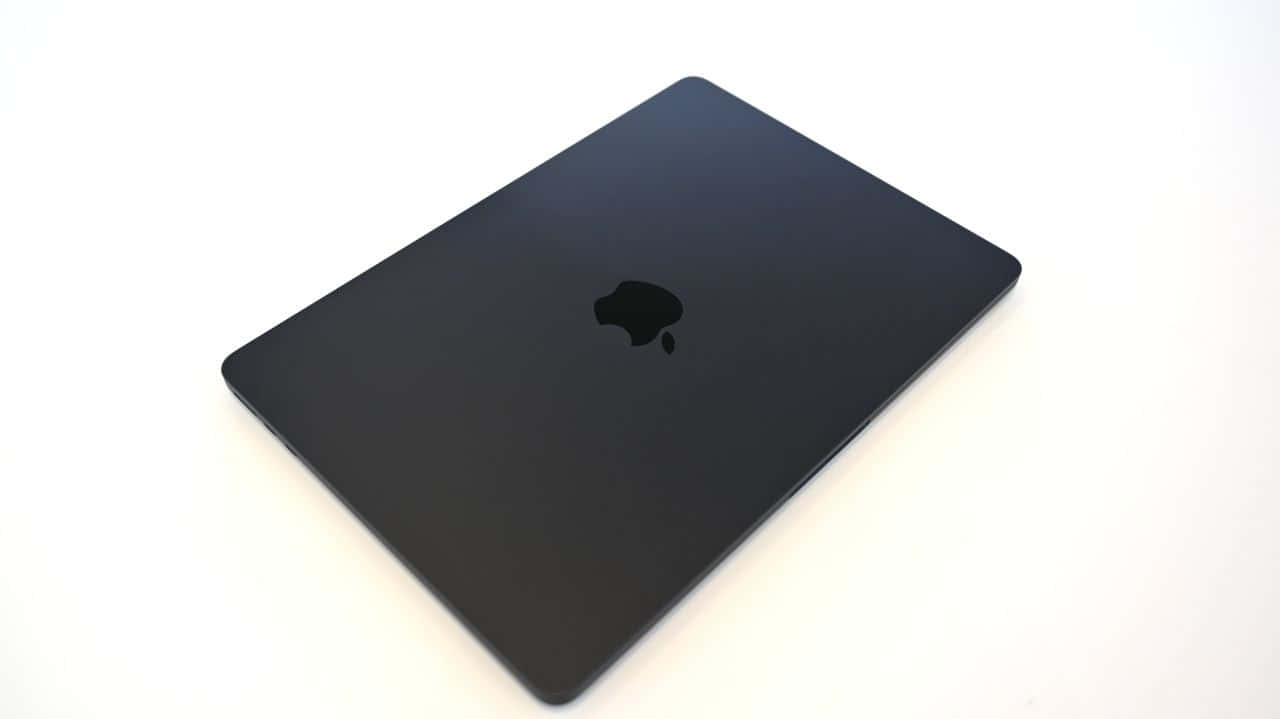 Black Macbook 桌布 壁紙 — 一個時尚的黑色MacBook蓋子以傾斜角度放置在純白表面上，其標誌性標誌隱約可見。