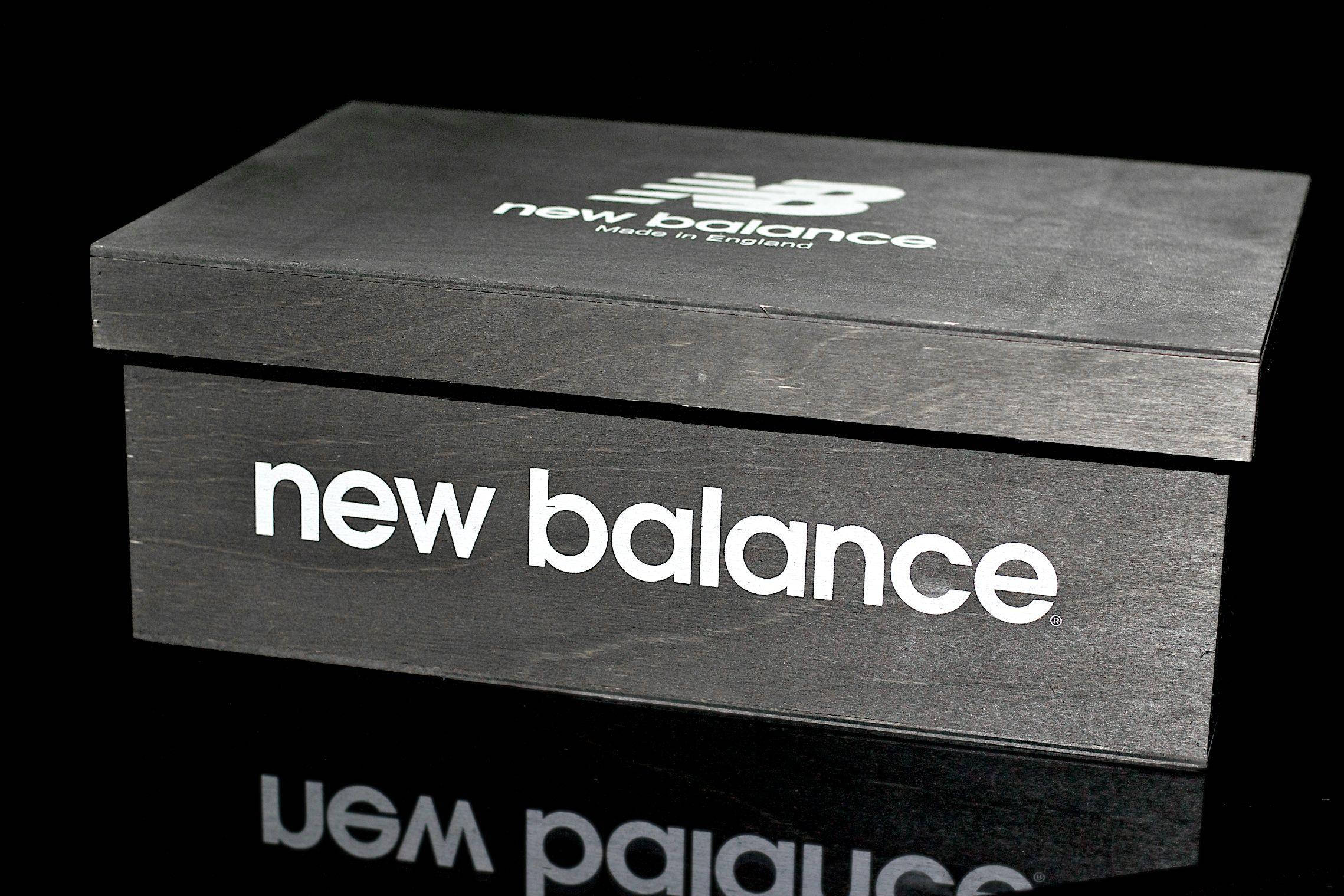 New 桌布 壁紙 — 一個時尚的黑色New Balance鞋盒，帶有白色品牌標誌，放置在深色背景的反射表面上。