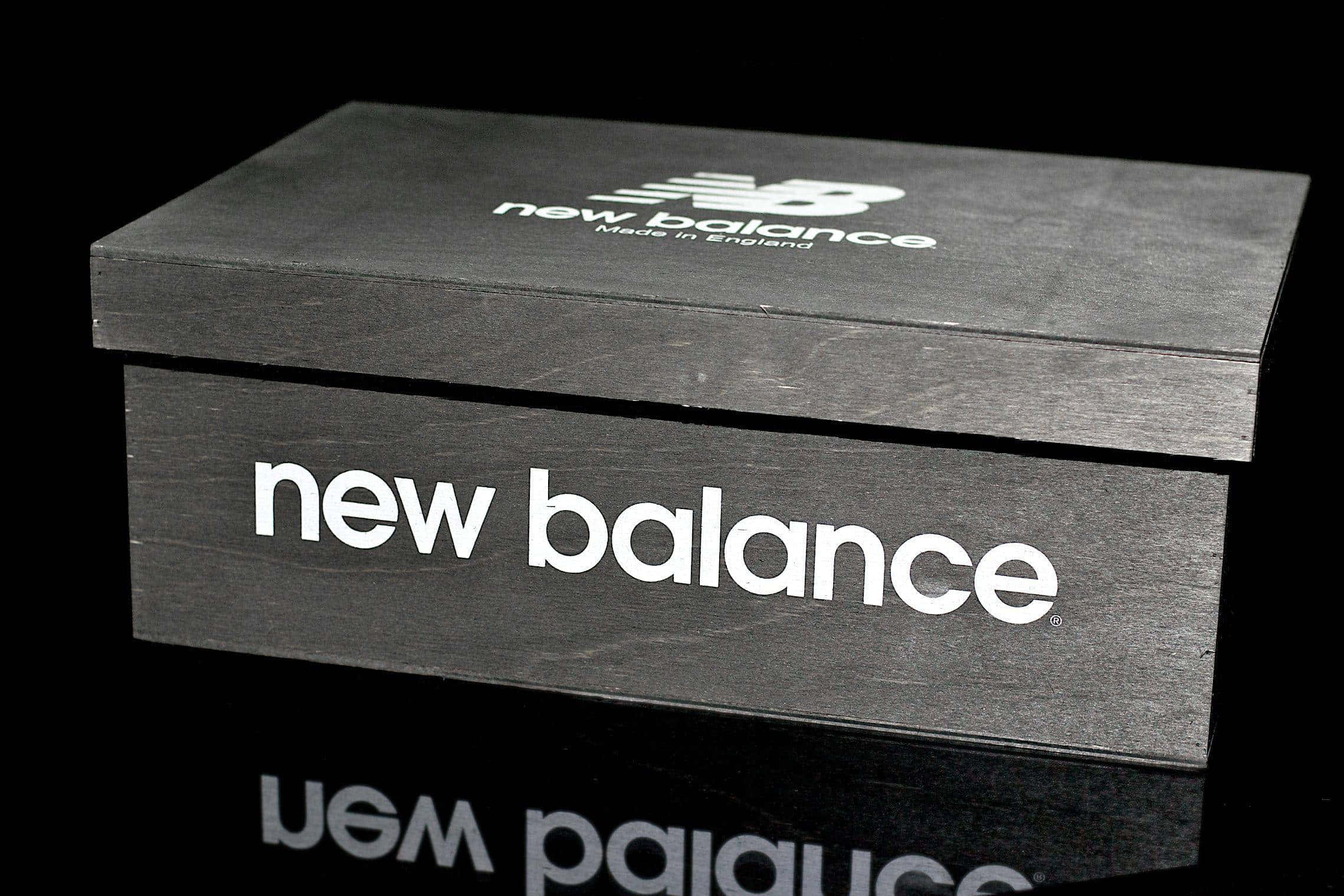 Shoe Box 桌布 壁紙 — 一個時尚的黑色New Balance鞋盒，帶有白色品牌標誌，放置在深色背景的反射表面上。