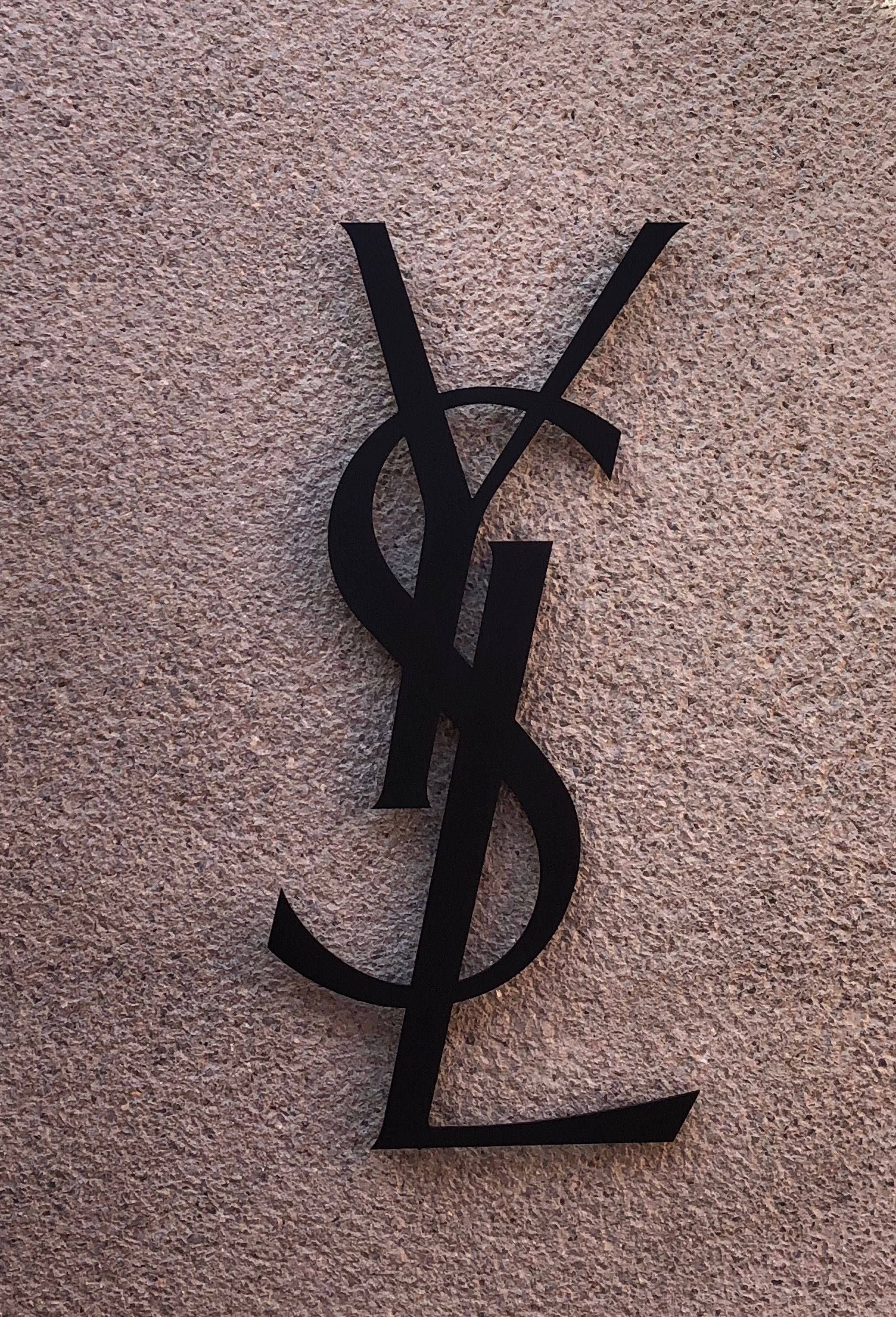 YSL 桌布 壁紙 — 一個時尚的黑色YSL字母組合標誌安裝在帶有細微紋理的米色牆上。
