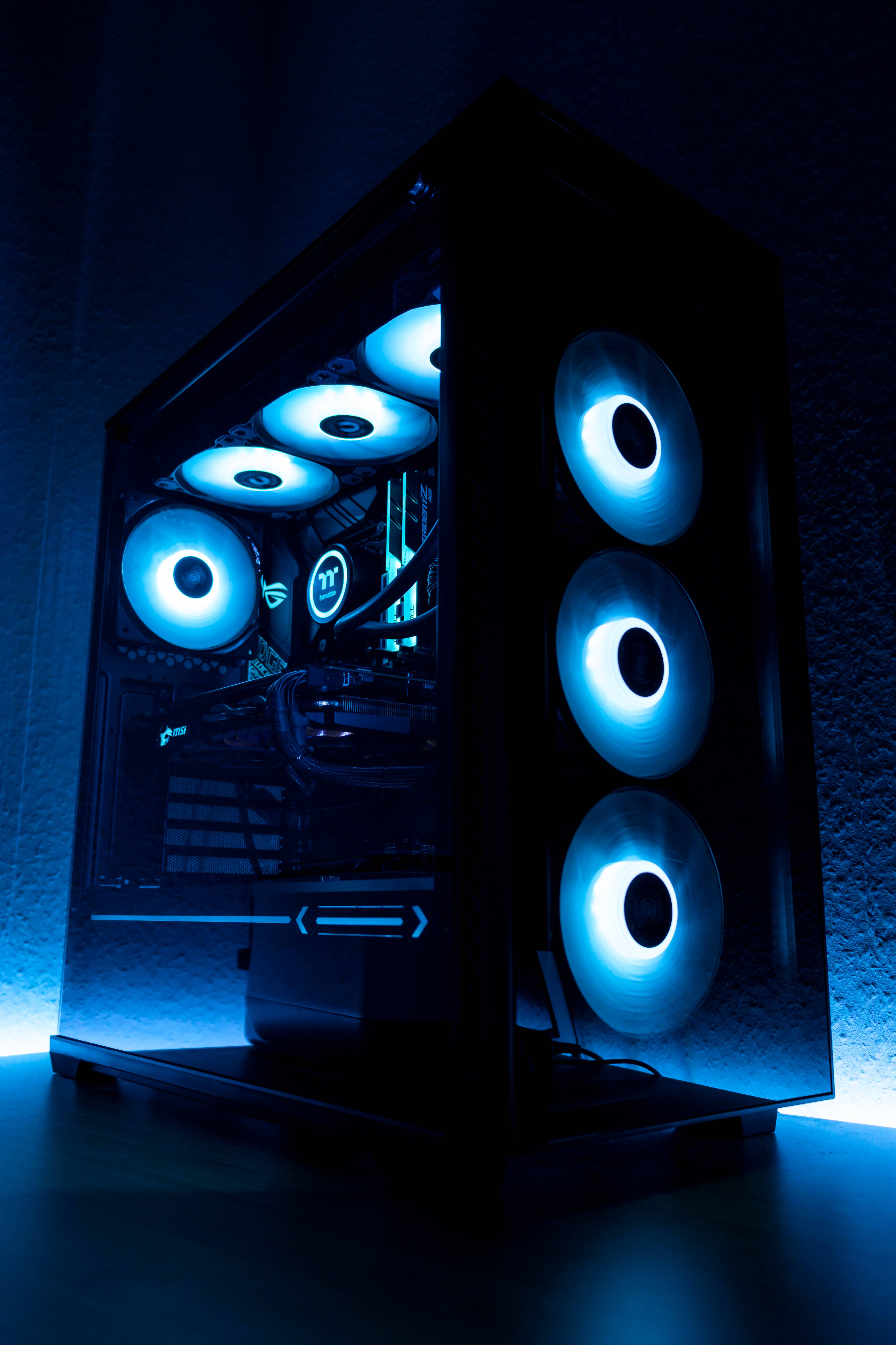 msi 2560x1080 桌布 壁紙 — 一個時尚的黑色電腦機殼，帶有發光藍色風扇和內部照明，靠在一面深色紋理牆上。