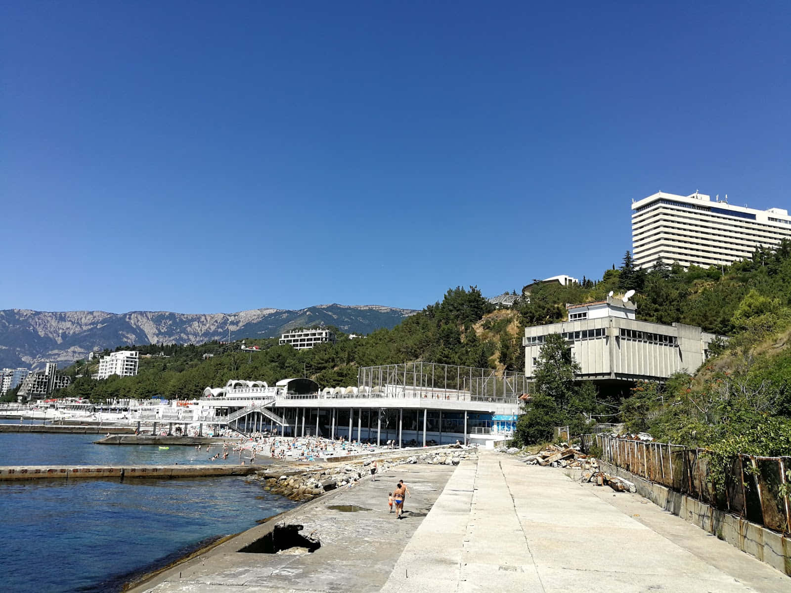 Boulevard 桌布 壁紙 — 一個晴朗的日子，在一個沿海小鎮，人們在碧綠的海水和山脈背景下，沿著混凝土碼頭游泳和散步。