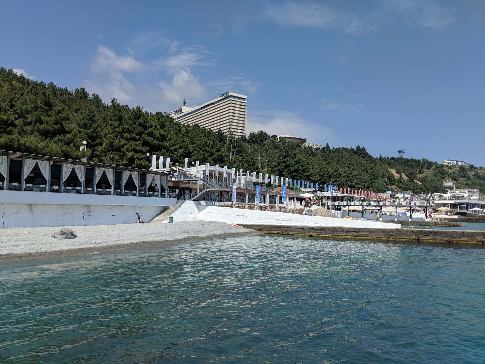 Port 桌布 壁紙 — 一個晴朗的日子，在海岸度假勝地，有碧綠的海水、溫柔的海浪、白色的卵石海灘和一座現代化酒店，坐落在綠色山丘之中。