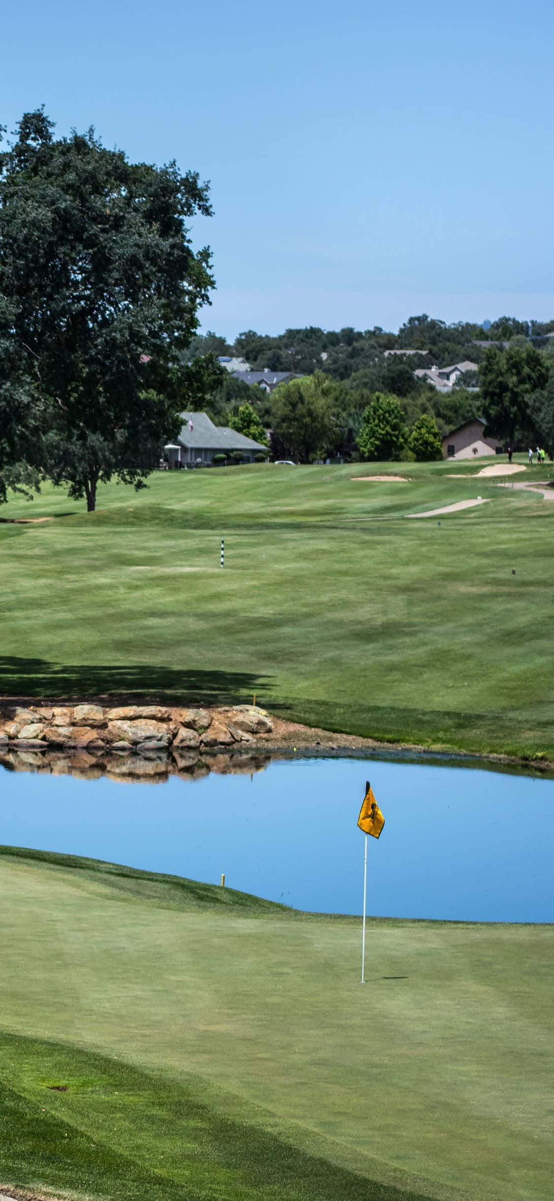 iPhone X Golf Course 桌布 壁紙 — 一個晴朗的日子，在鬱鬱蔥蔥的高爾夫球場上，黃色旗幟標示著靠近平靜水障礙的球洞。