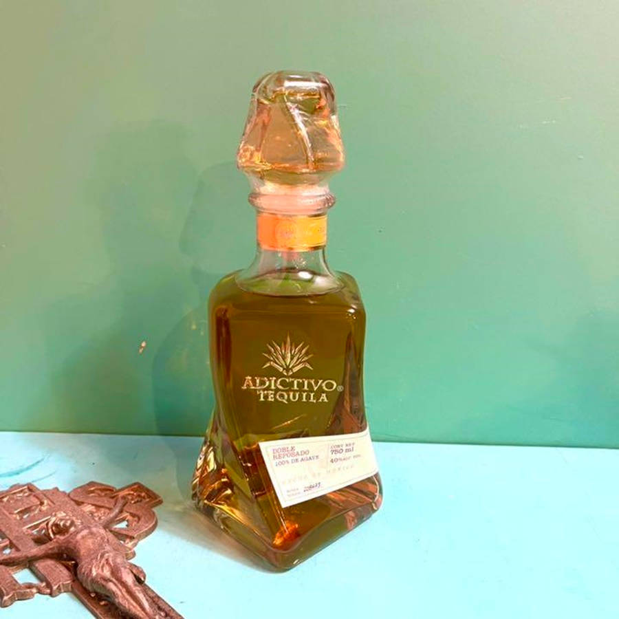筆電 桌布 壁紙 — 一個有切割面的Adictivo Tequila酒瓶放在青綠色表面上，旁邊有一個青銅十字架，背景是薄荷綠色的牆壁。
