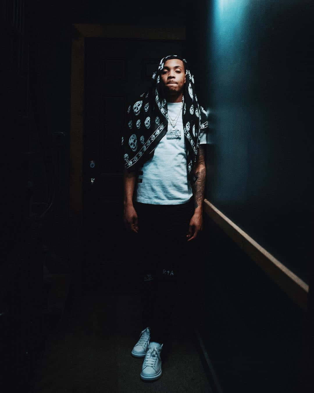 Herbo iPhone 桌布 壁紙 — 一個有刺青、戴著骷髏圖案頭巾的年輕男子站在昏暗的走廊中，被頭頂上的一盞燈照亮。