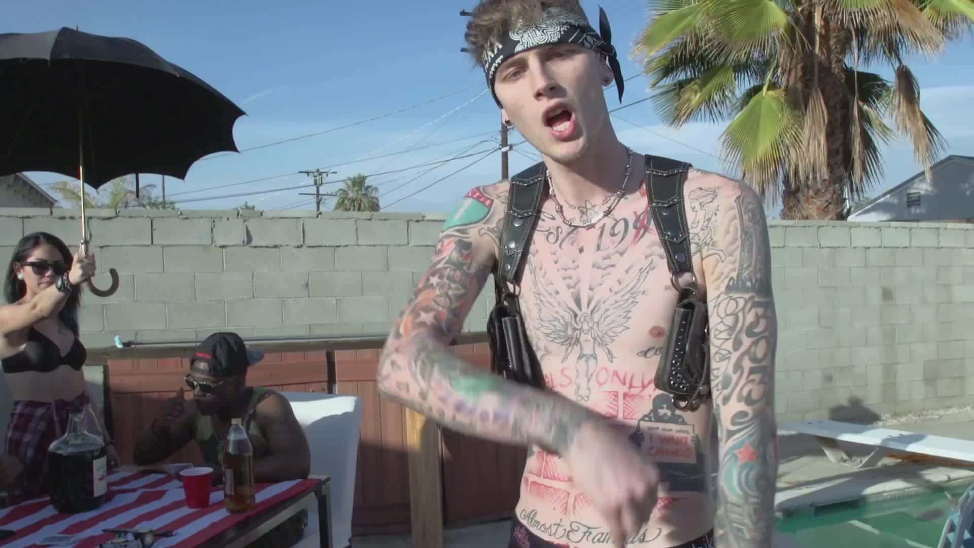 mgk 桌布 壁紙 — 一個有刺青、戴頭巾的男人在戶外熱情地唱歌，朋友們在泳池旁的黑傘下放鬆。