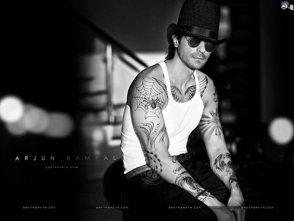 Arjun Rampal 桌布 壁紙 — 一個有刺青、戴高帽的男人自信地坐在昏暗的城市環境中，背景有模糊的燈光。