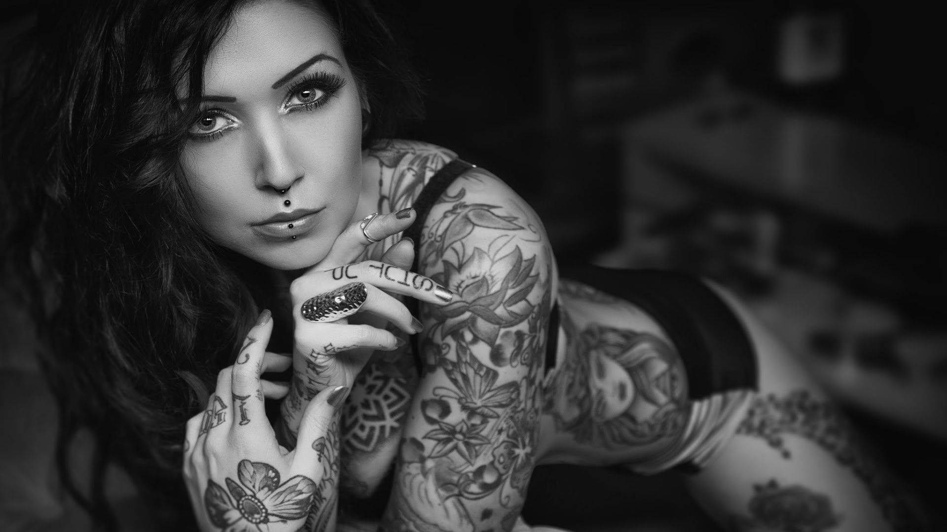 Tattoo 桌布 壁紙 — 一個有刺青、深色頭髮和銳利眼神的女人，在陰鬱的黑白肖像中直視鏡頭。