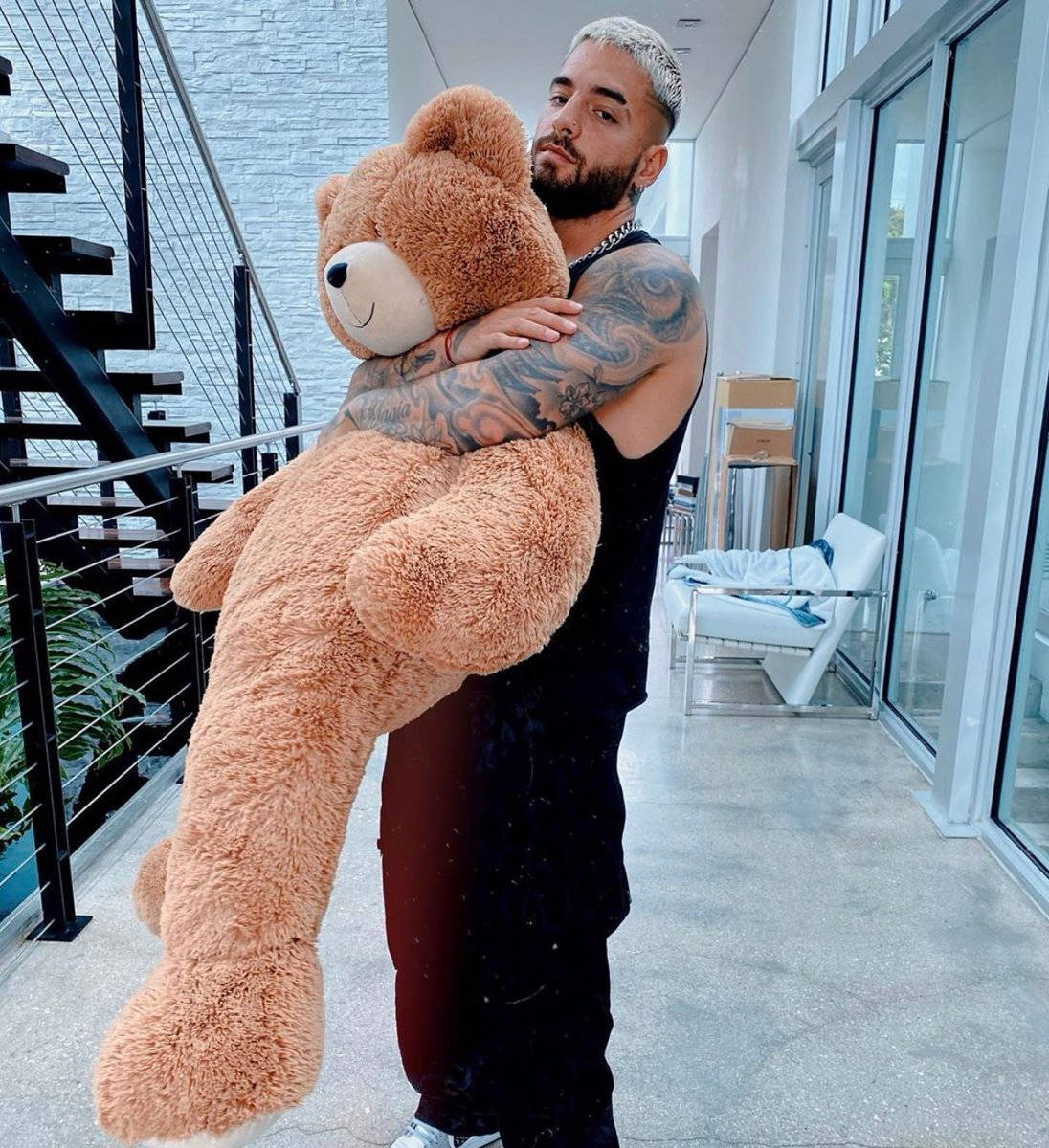 Maluma 桌布 壁紙 — 一個有刺青、漂染頭髮的男人在現代玻璃牆走廊抱著一個巨大的毛茸茸棕色泰迪熊。