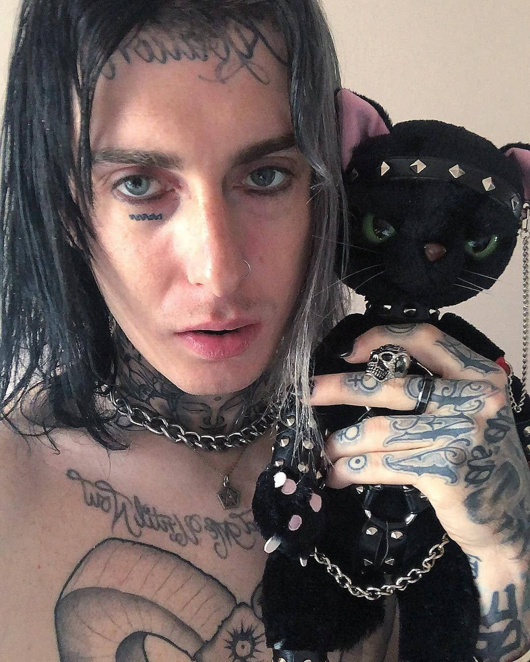 Ghostemane 桌布 壁紙 — 一個有刺青、皮膚蒼白、深色頭髮的人，拿著一隻黑色鉚釘貓咪絨毛玩具，靠著一面素牆。