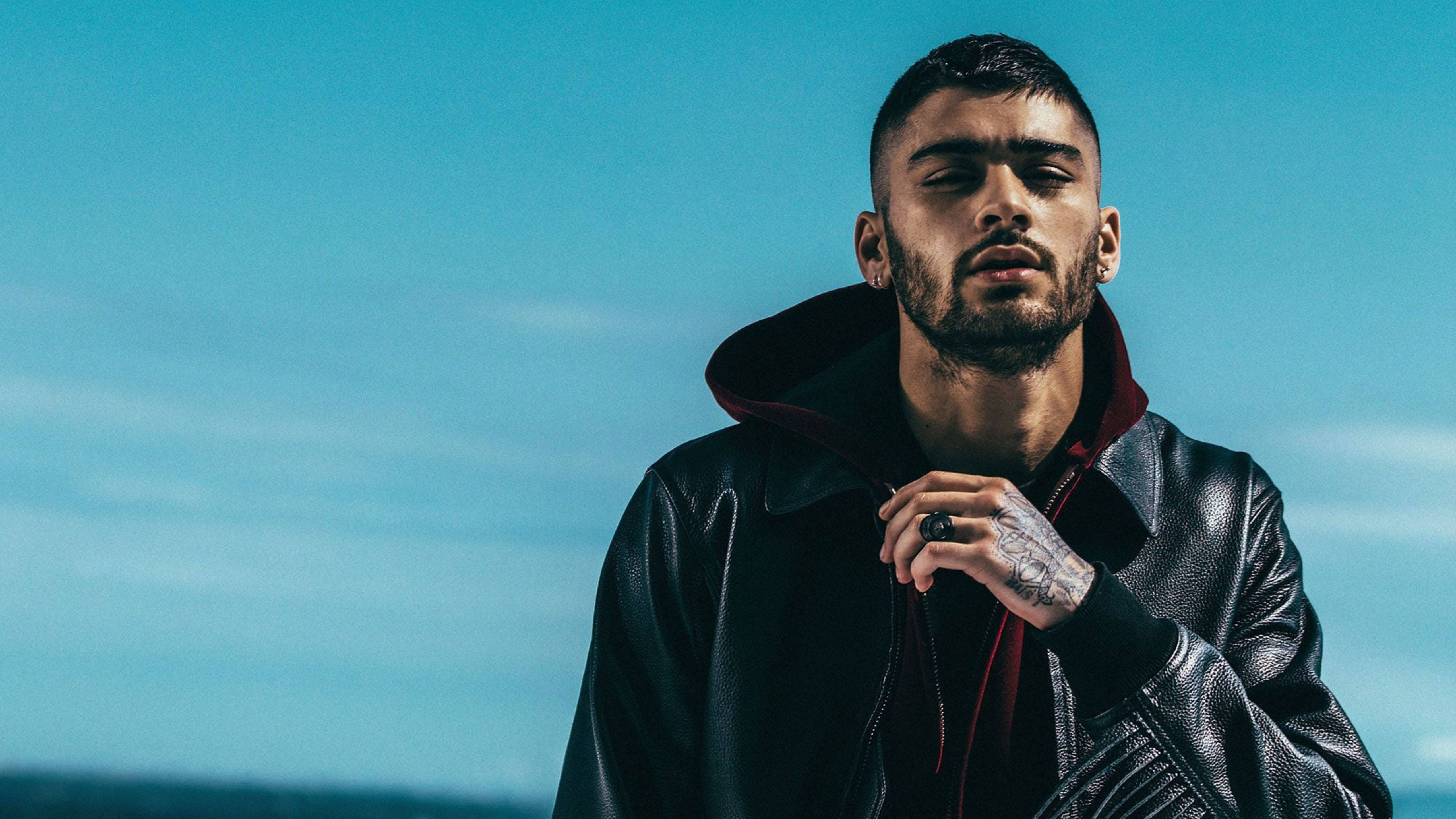 zayn 桌布 壁紙 — 一個有刺青、穿著皮夾克的男人，以嚴肅的表情站在清澈的藍天下。