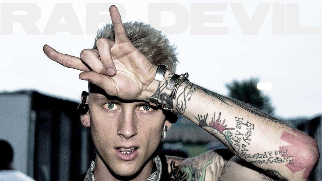 mgk 桌布 壁紙 — 一個有刺青、綠眼睛和金色頭髮的男人，一邊做出手勢，一邊專注地看著鏡頭。