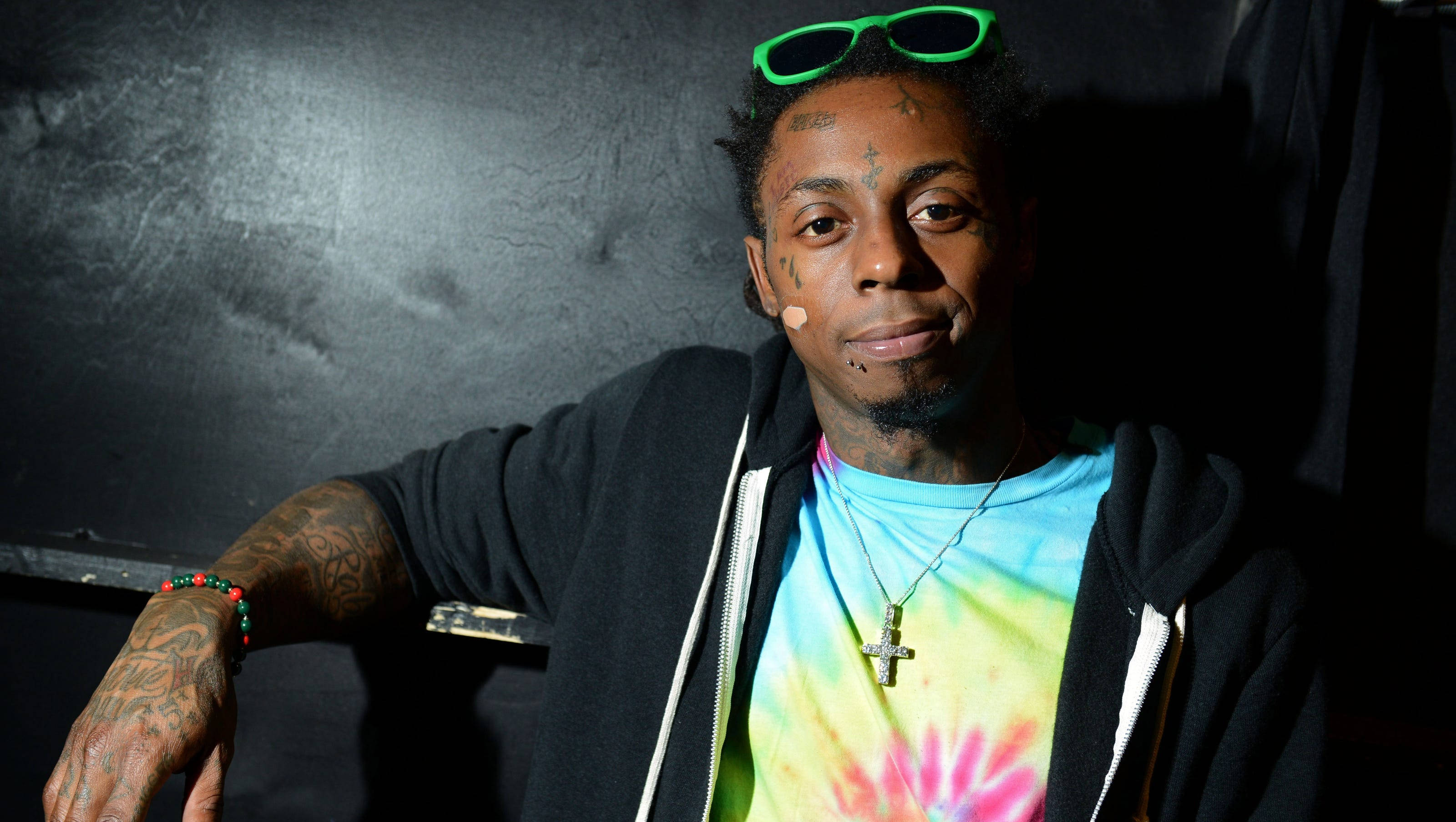 Lil Wayne 桌布 壁紙 — 一個有刺青、頭上戴著綠色太陽眼鏡的男人，在深色紋理牆壁前穿著黑色連帽衫下的紮染襯衫。